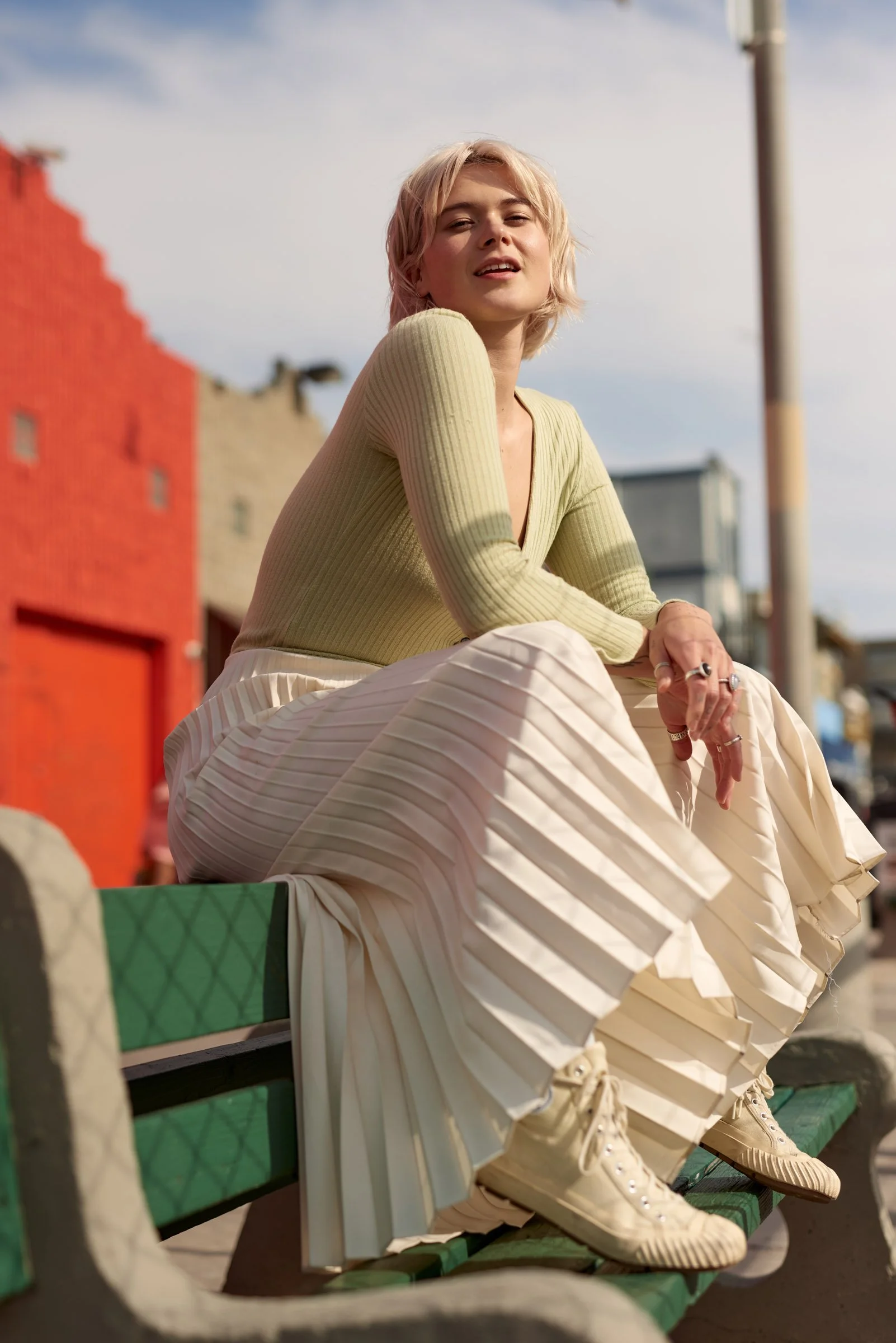 Young woman with blonde hair sitting on a green park bench outdoors, wearing a beige pleated skirt, a light yellow ribbed sweater, and matching sneakers, with a cityscape in the background.