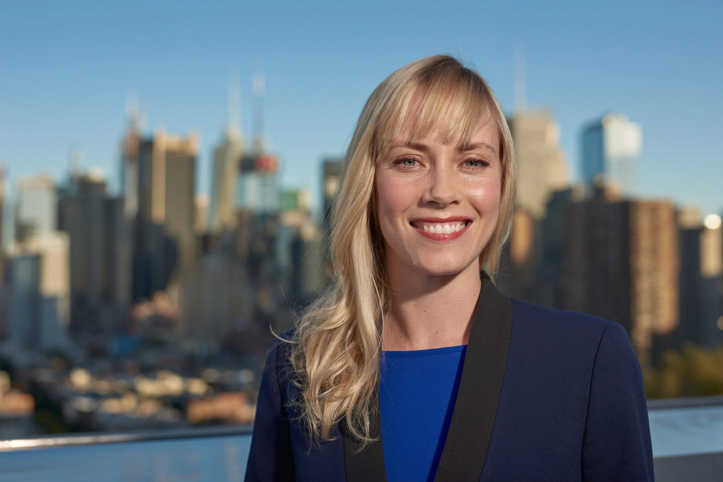 A smiling woman with blonde hair and a blue blazer, standing with a city skyline in the background.