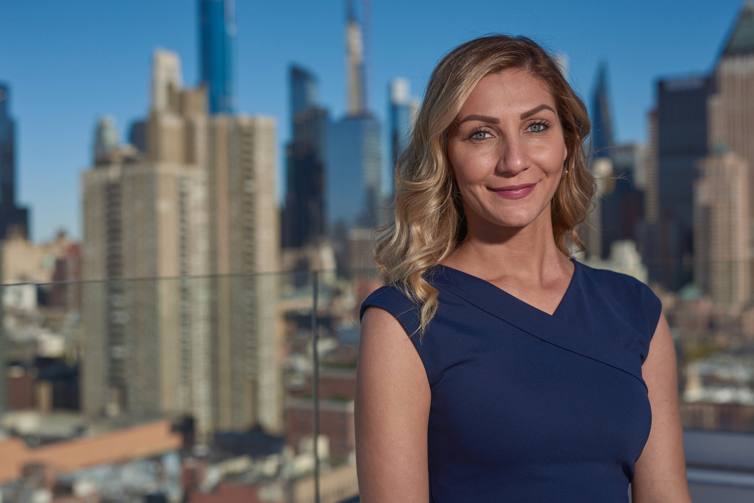A woman with blonde hair in loose waves, wearing a blue sleeveless top, standing outdoors with a city skyline in the background on a clear day.