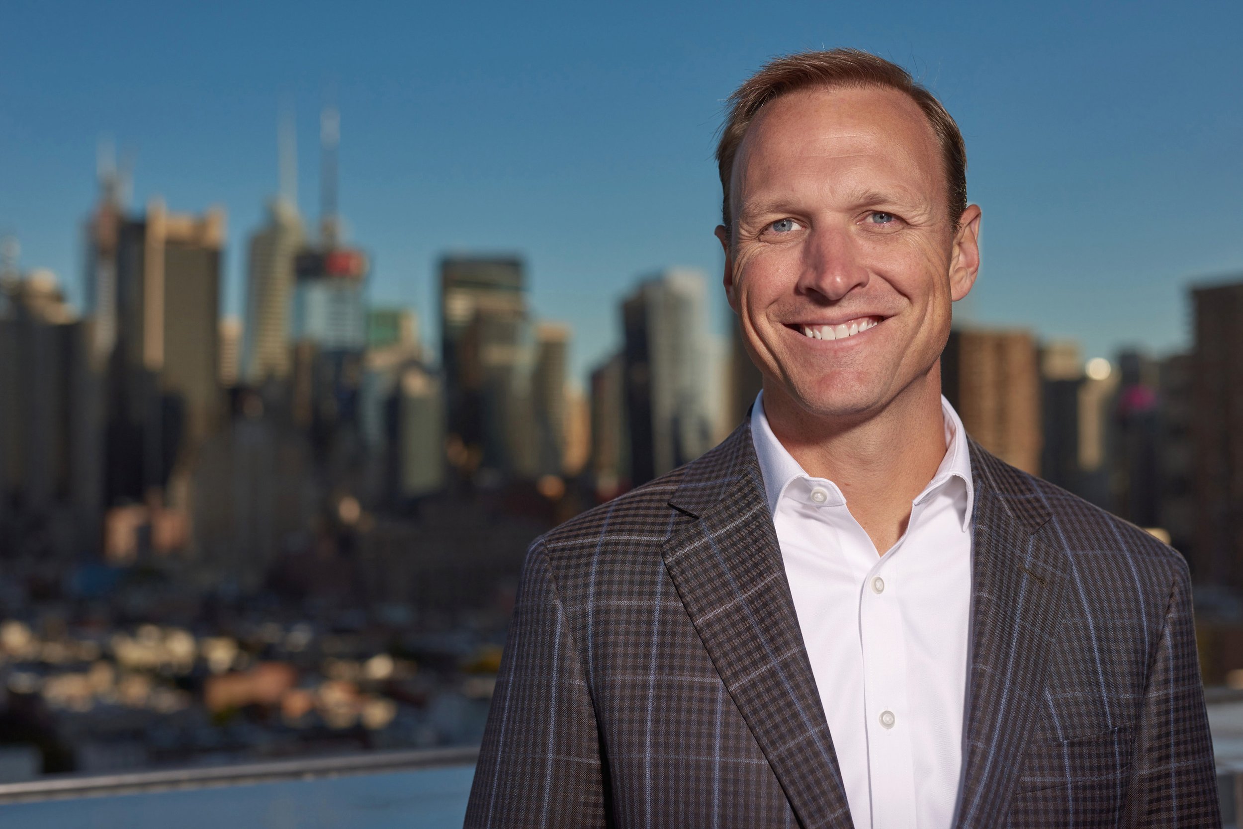 A man wearing a checkered blazer and white shirt smiling in front of a city skyline with tall buildings and a clear blue sky.