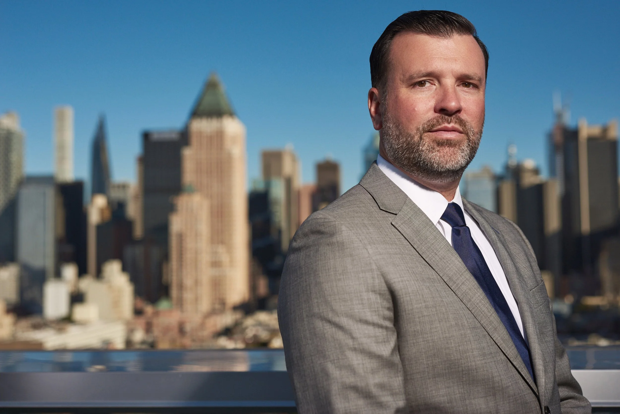 A man in a grey suit with a blue tie on a rooftop with a city skyline in the background.