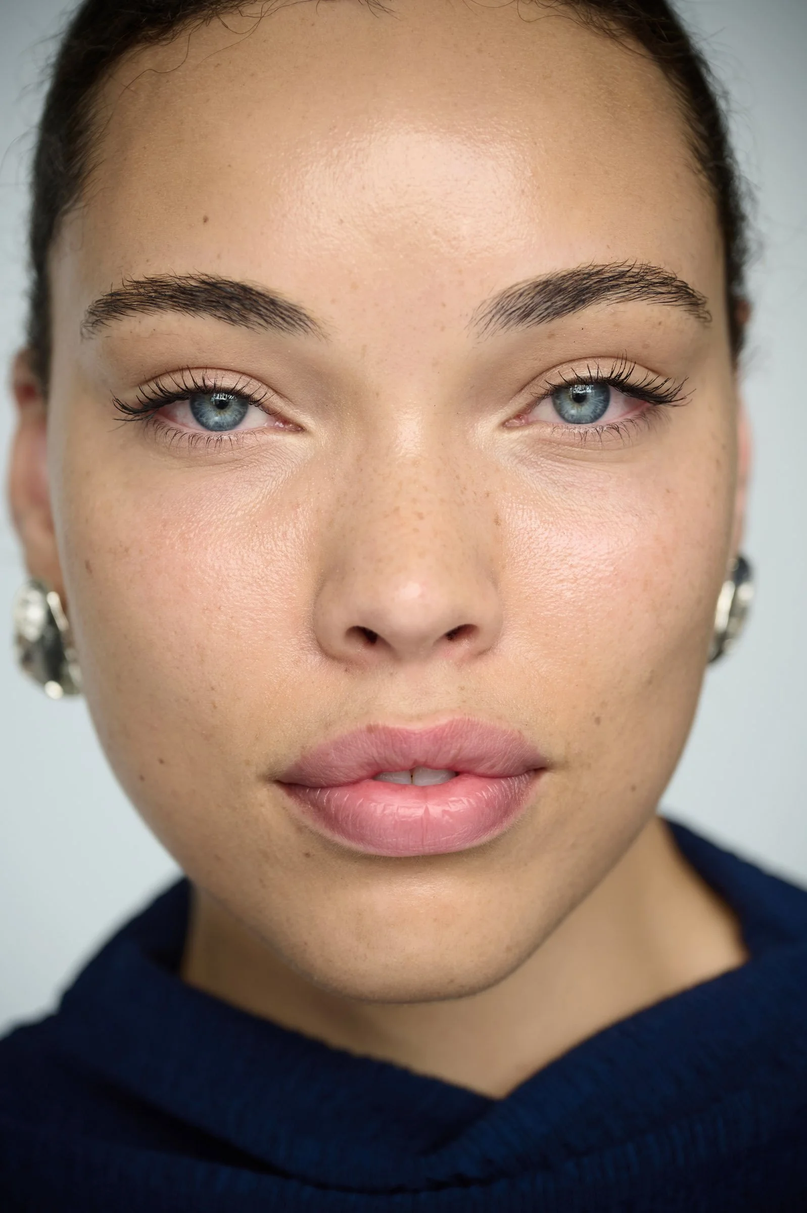 Close-up of a woman with blue eyes, dark eyebrows, light makeup, wearing earrings and a dark top.