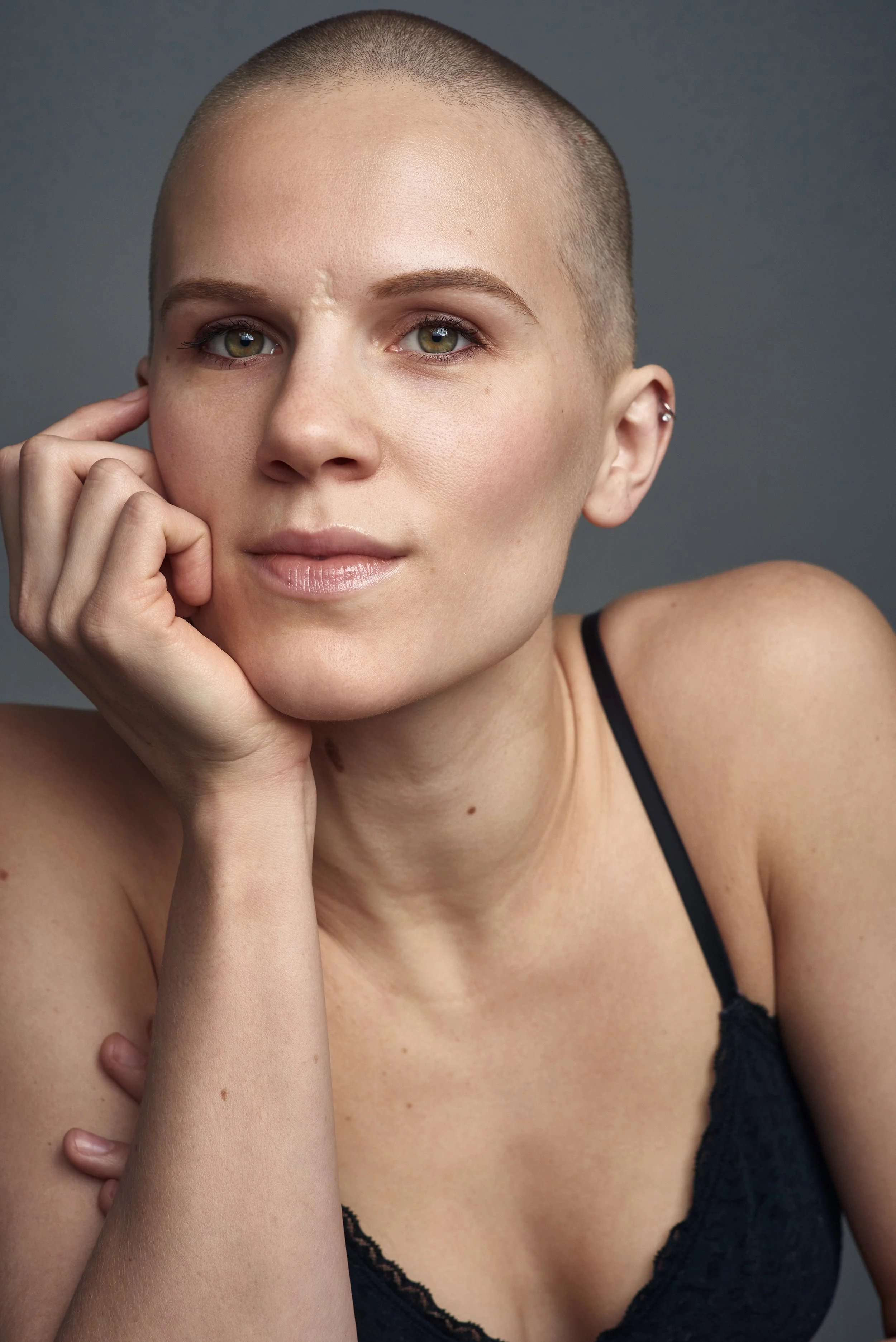 A woman with a shaved head wearing a black lace top, resting her chin on her hand, looking directly at the camera against a gray background.