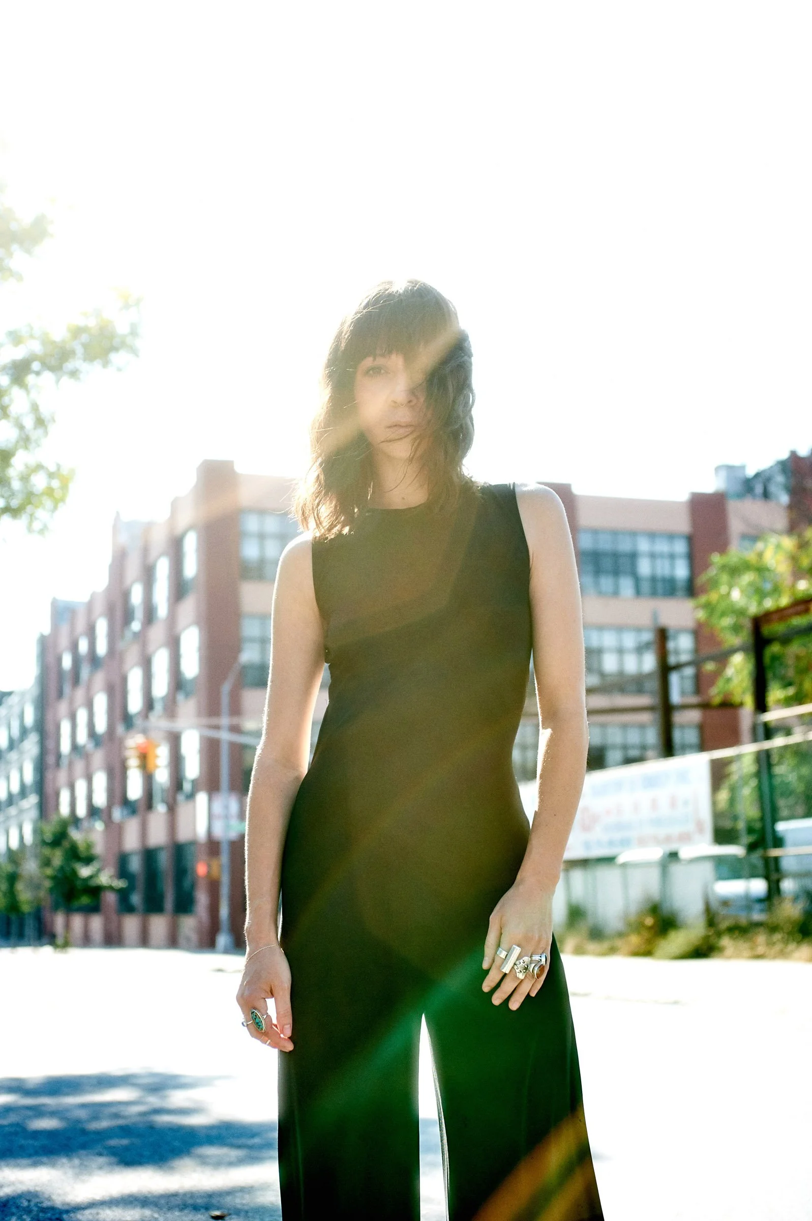 A woman standing outdoors in front of sunlight, wearing a sleeveless black jumpsuit, with buildings and trees in the background.