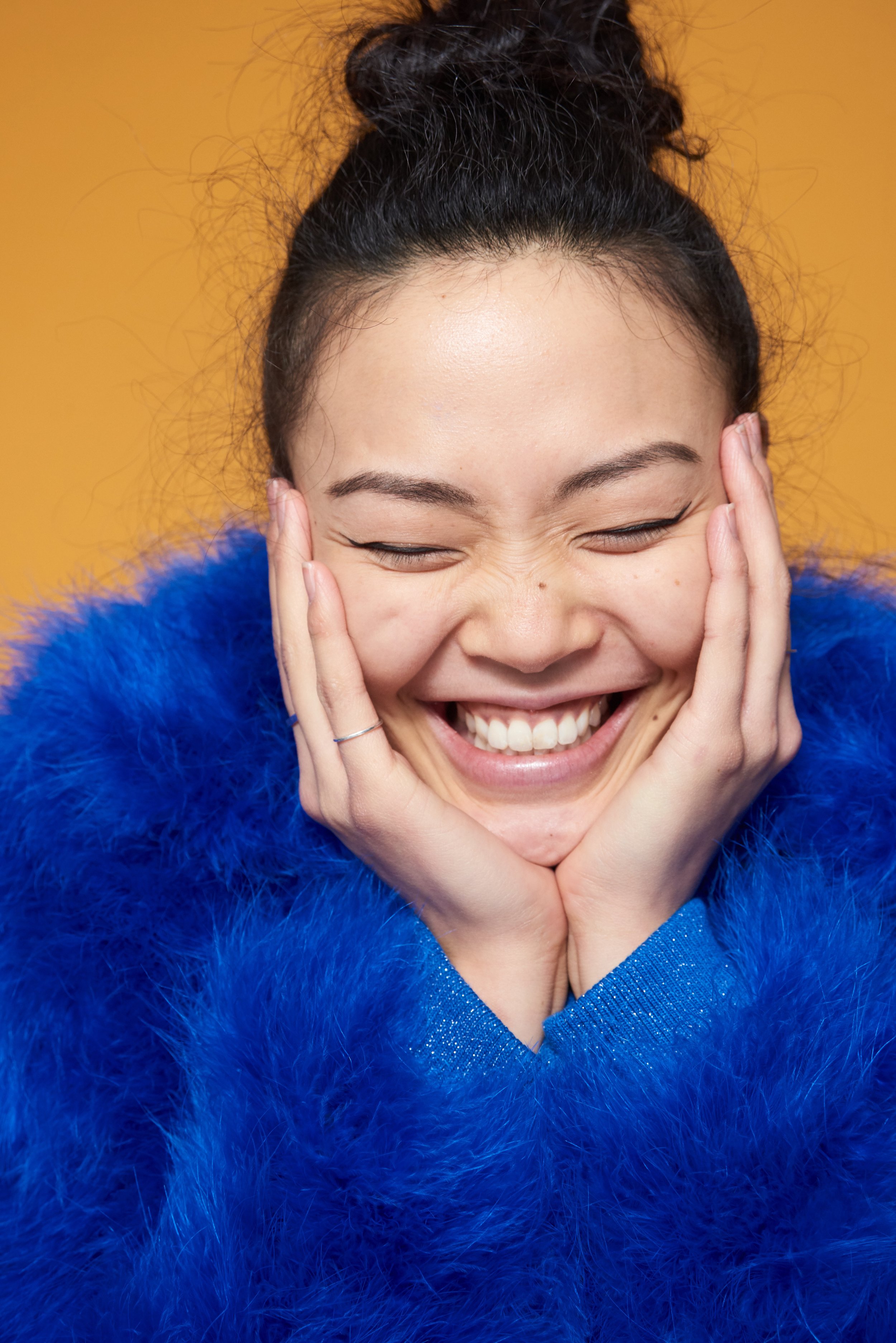 A woman with dark hair in a bun, smiling brightly with eyes closed, wearing a bright blue fuzzy sweater.