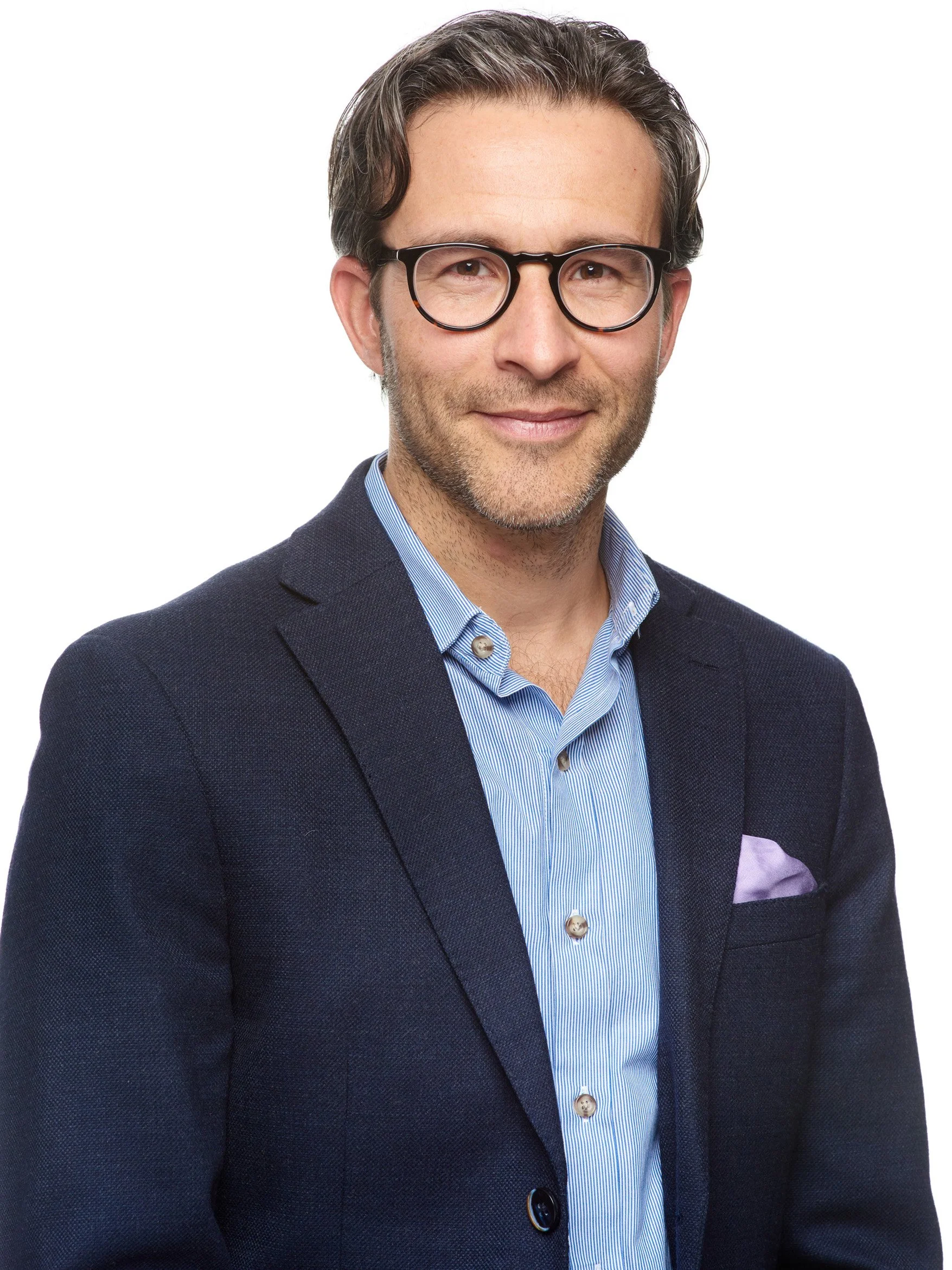 A man with dark hair, glasses, and a light beard wearing a dark blazer and a striped blue shirt, smiling at the camera against a white background.