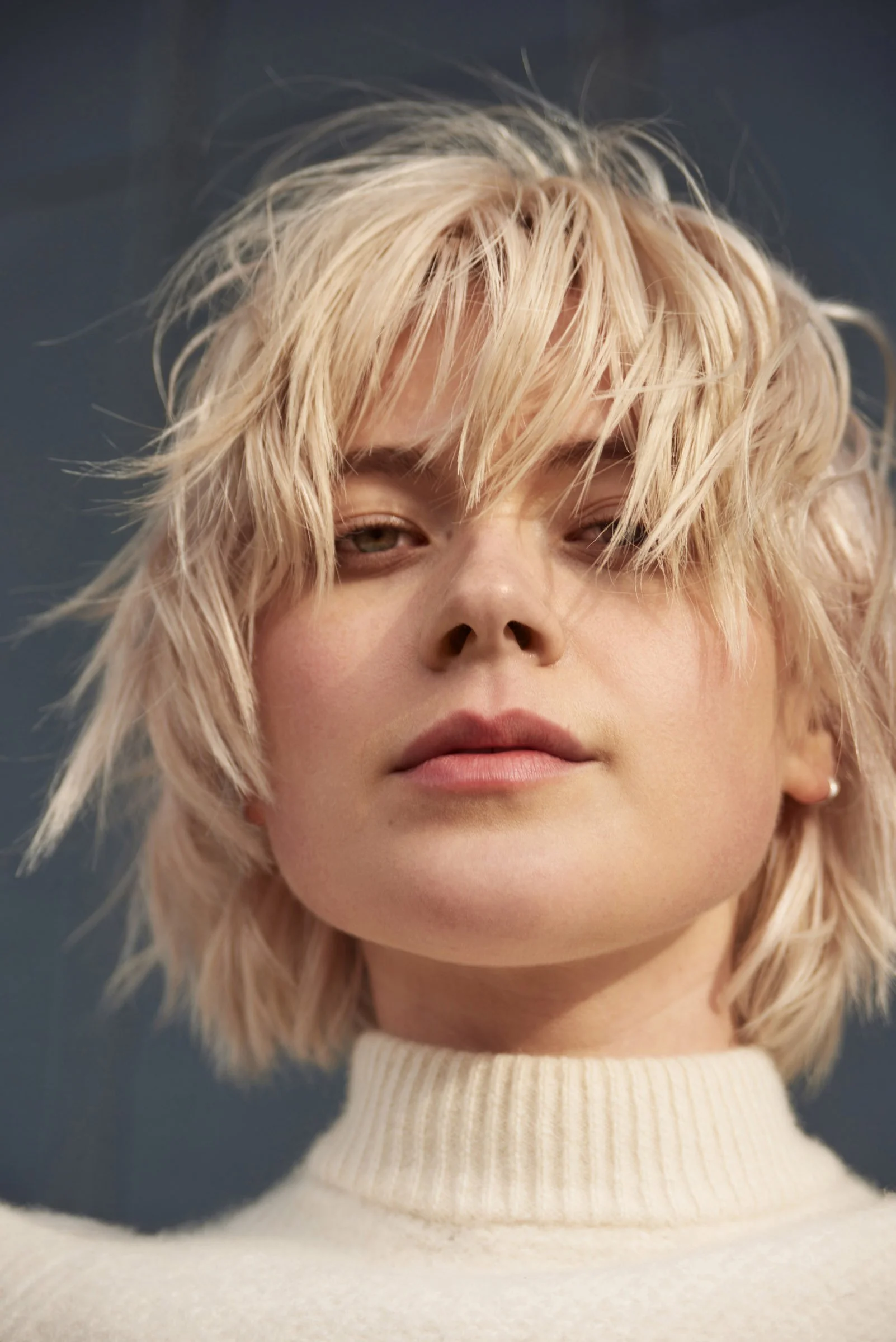 A woman with messy blonde hair, partially covering her face, wearing a white turtleneck sweater against a dark background.