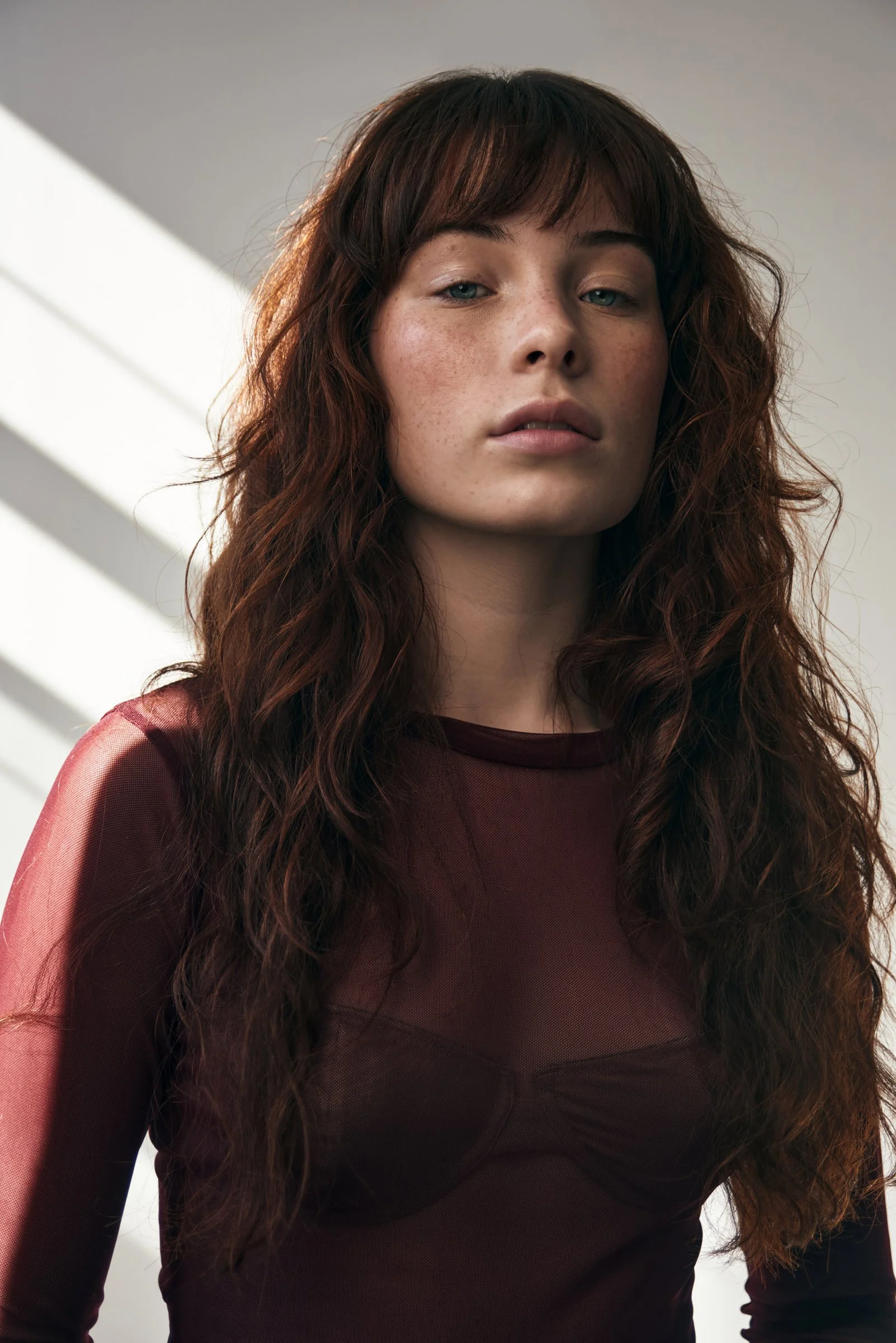 A young woman with long, wavy red hair and freckles wearing a sheer, long-sleeved maroon top, standing indoors with sunlight streaming through blinds in the background.