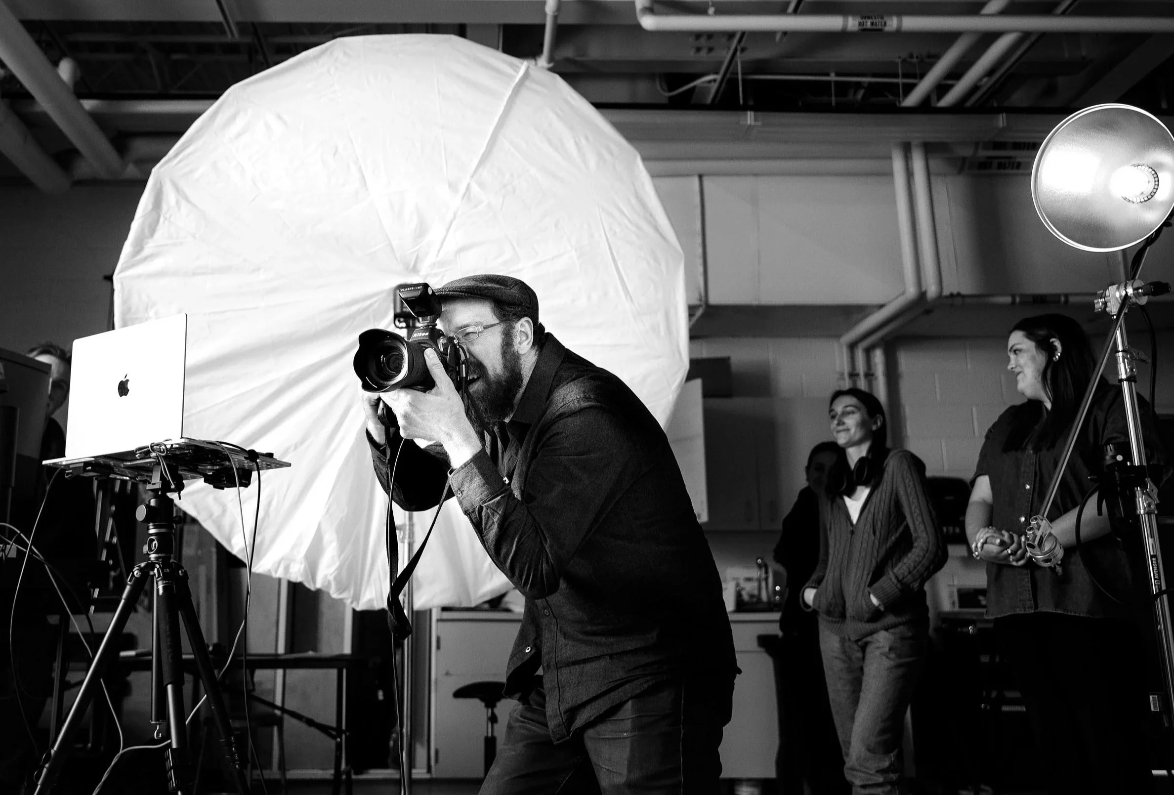 Photographer taking a picture in a studio with a large white umbrella and studio lights, while two women observe in the background.