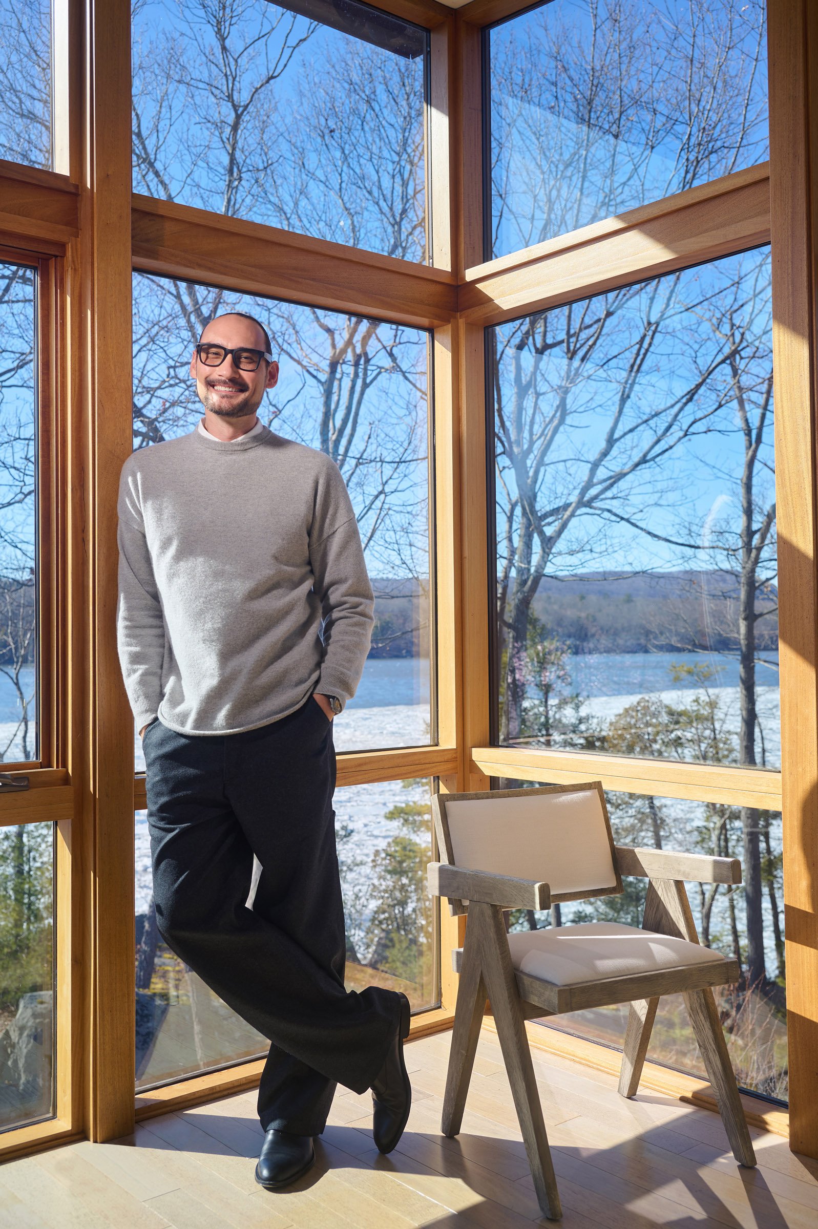 A man smiling and standing inside near large windows with a view of snow-covered trees and a lake.