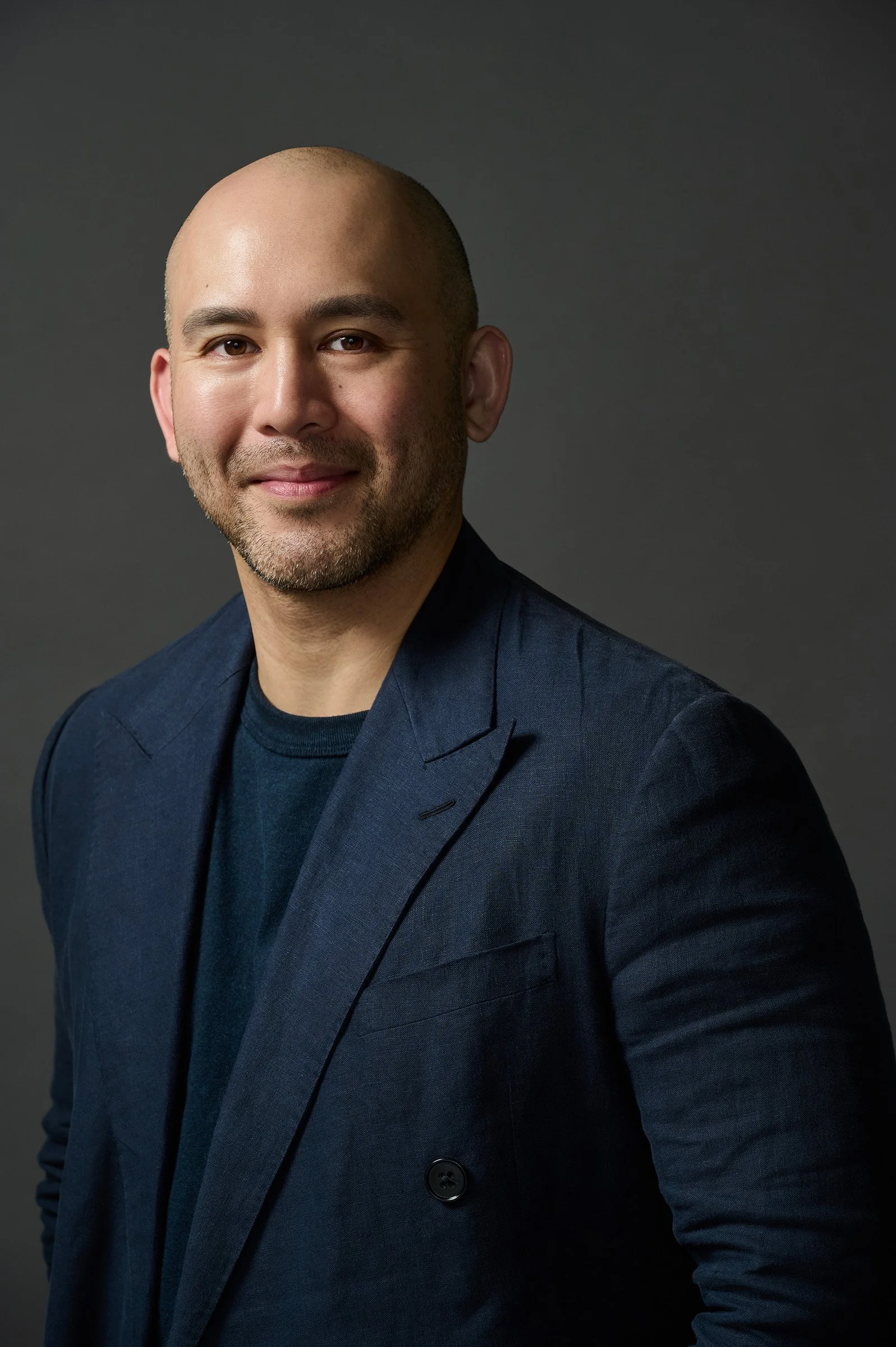 A man with a shaved head and beard wearing a dark blazer and shirt, posing against a dark gray background.