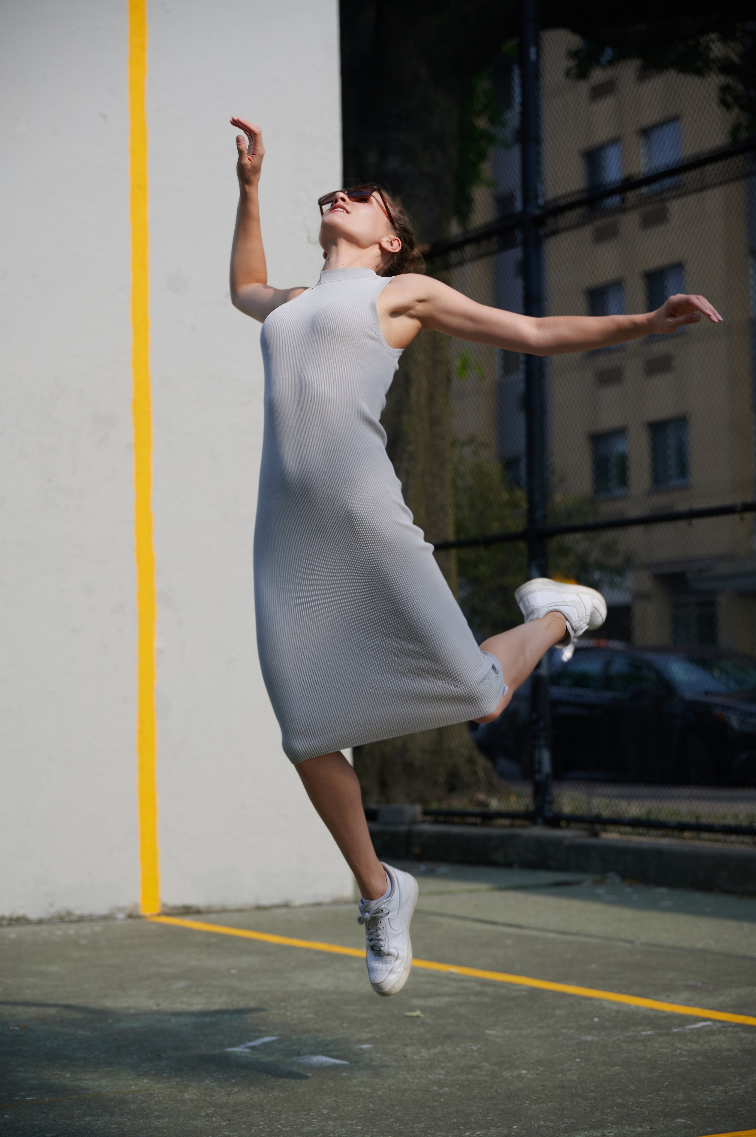 A woman in a gray sleeveless dress and white sneakers jumping in the air on a sports court with a white wall and black chain-link fence in the background.