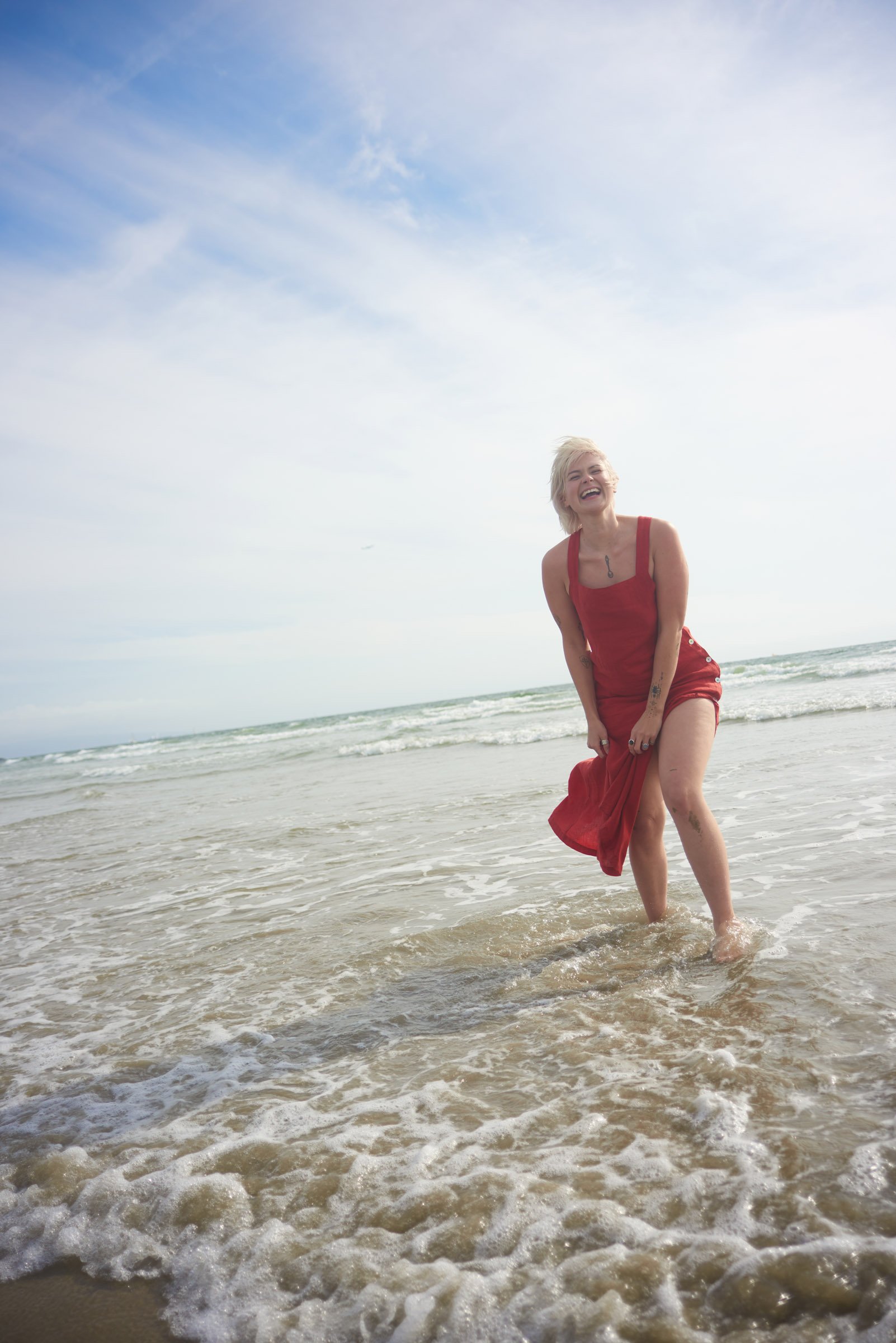 A woman with short blonde hair wearing a red dress, joyfully standing in shallow ocean water on a beach, smiling and laughing.