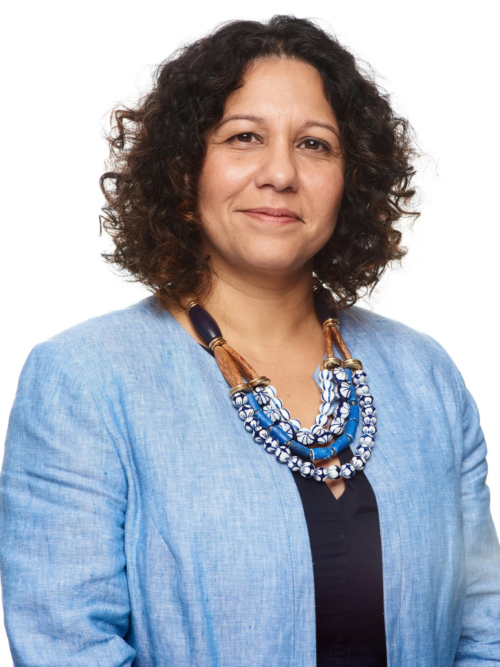 Headshot of a woman with curly dark hair, wearing a blue blazer over a black top and a multi-strand beaded necklace in blue, white, and brown tones, posing against a white background.