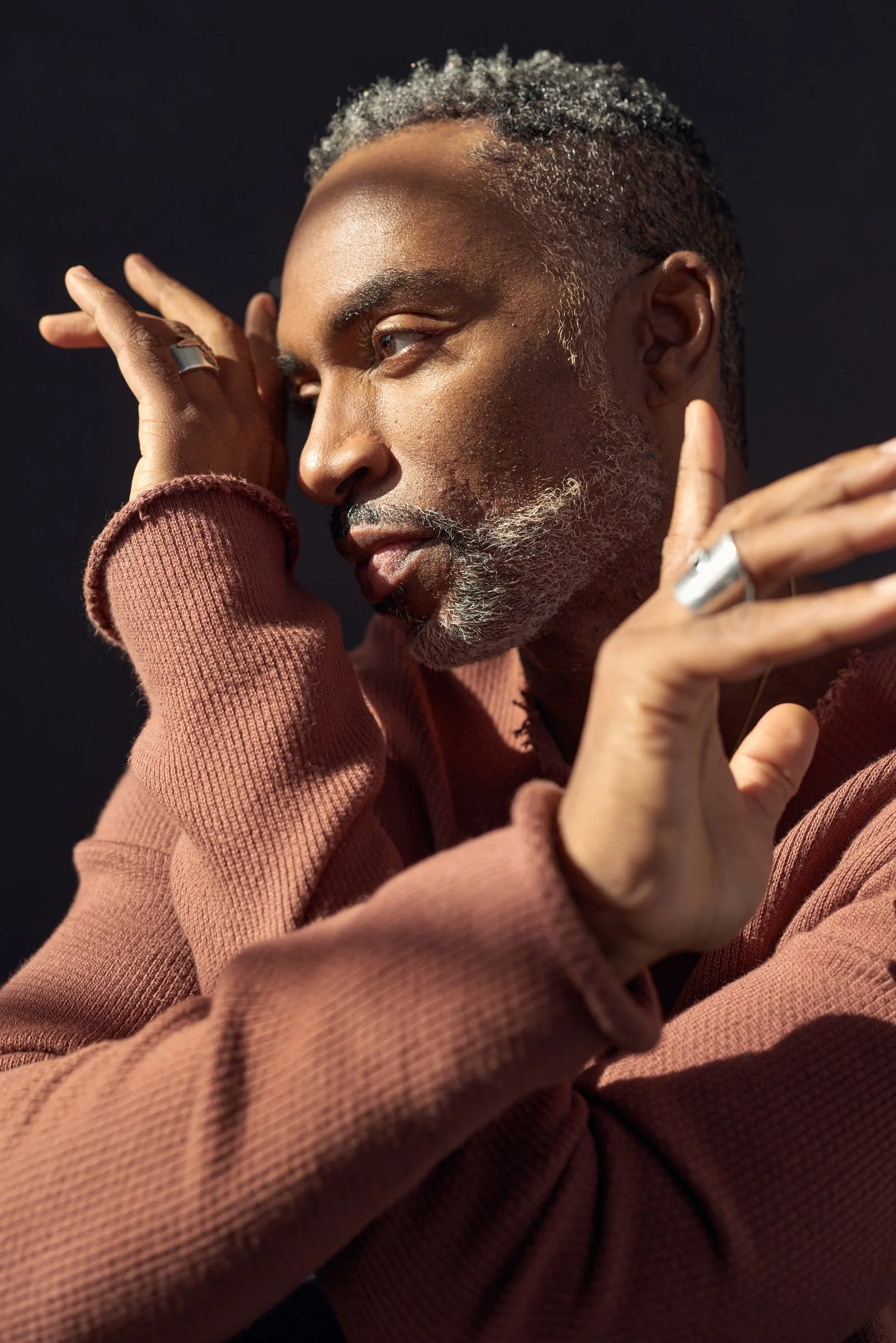 A side profile of a mature man with gray hair and beard, wearing a pinkish-brown sweater, posing with hands near his face against a dark background.