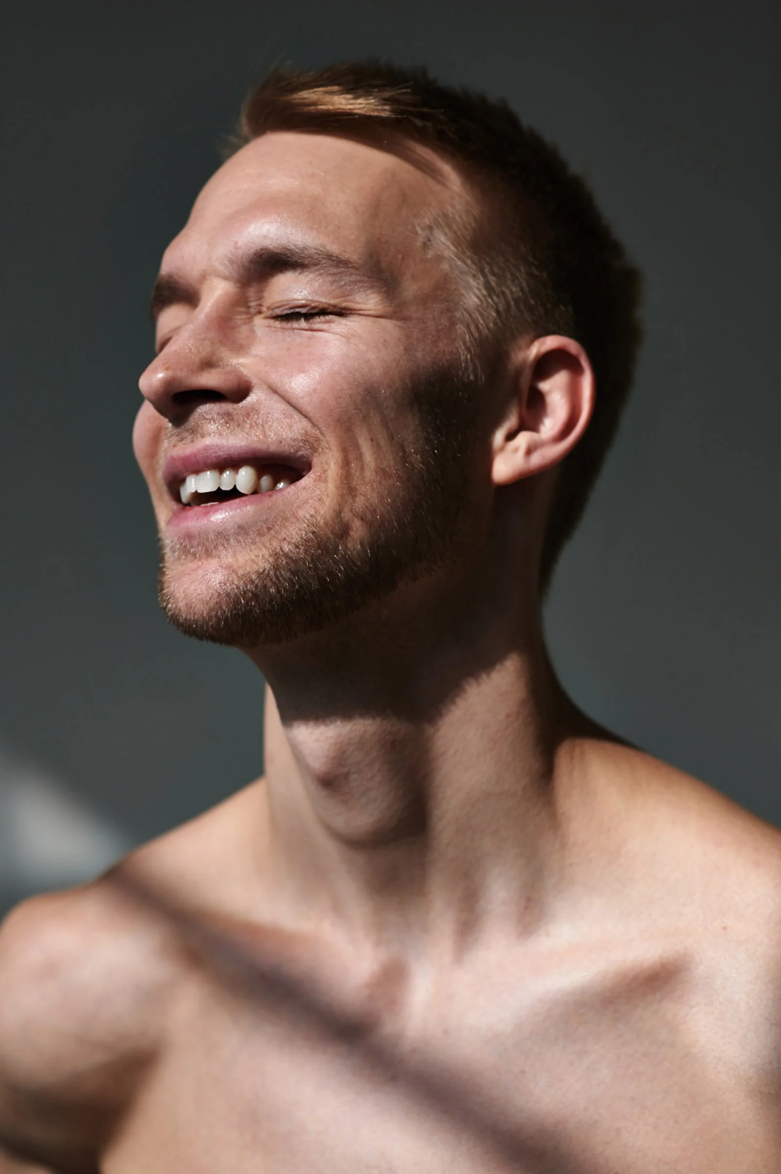 A shirtless man with short brown hair, a beard, and a big smile with eyes closed, against a dark background.