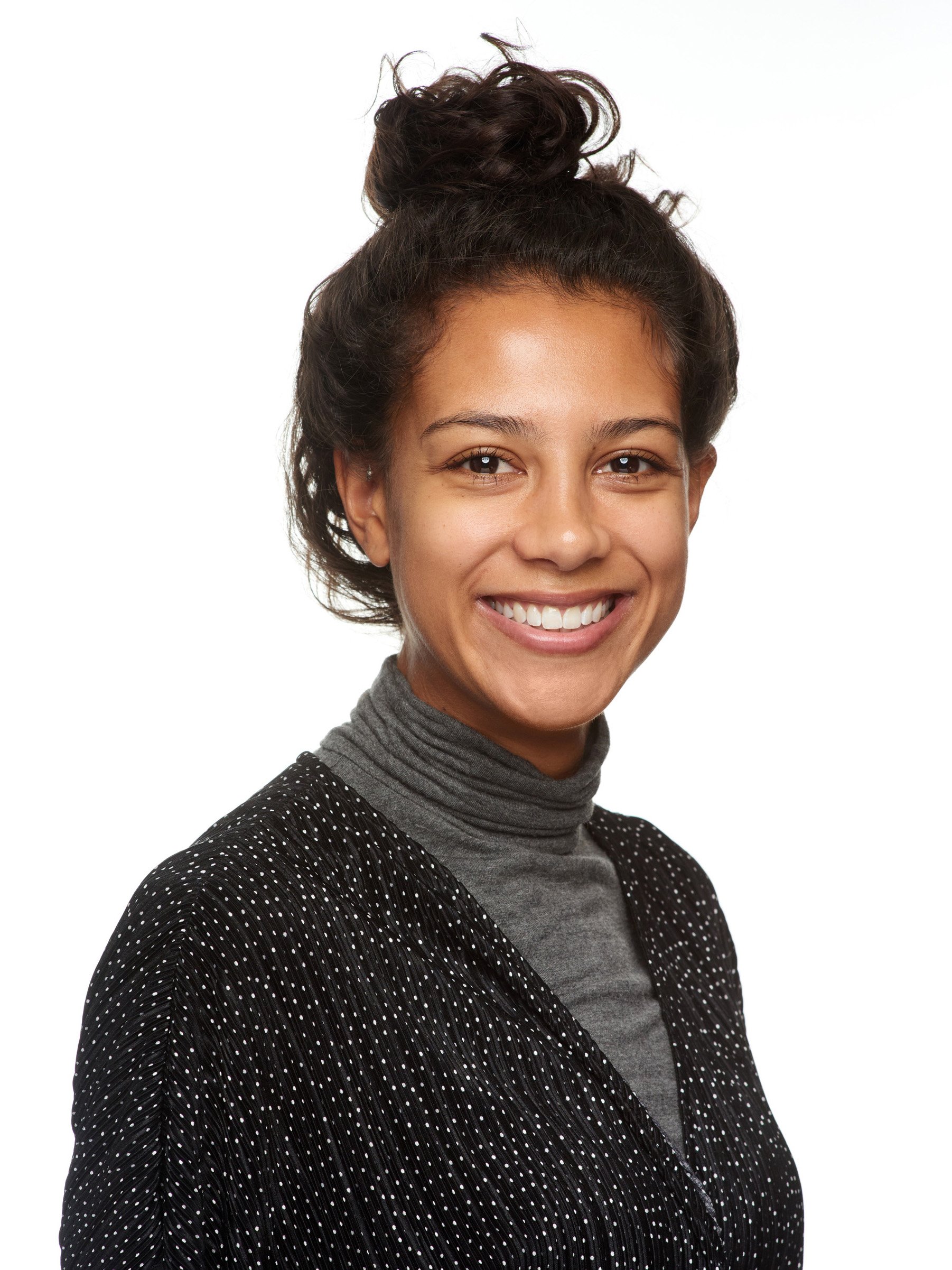 A young woman with dark hair styled in a messy bun, smiling, wearing a gray turtleneck and a black, polka-dotted blazer.
