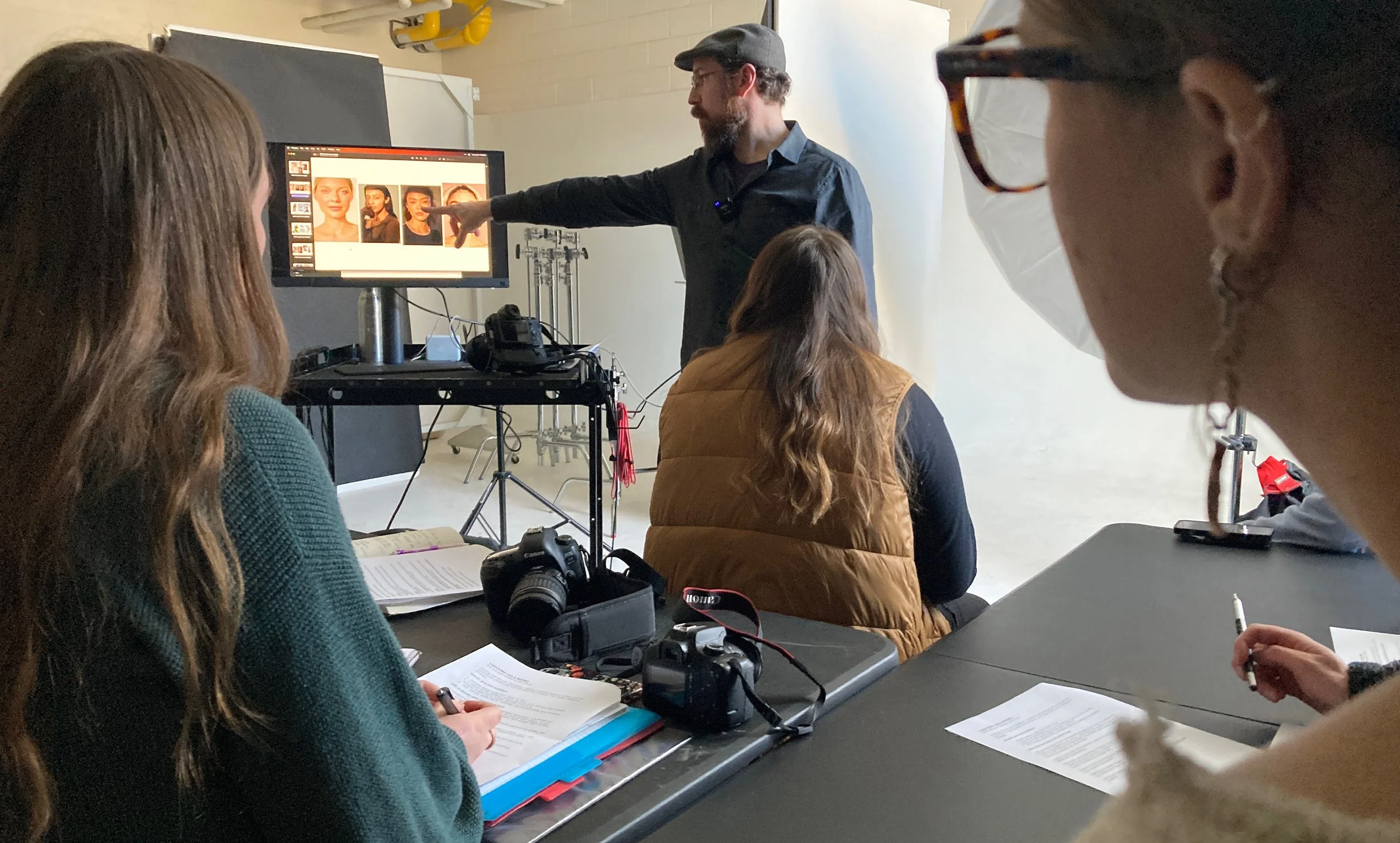 A photography class in a studio with students listening to an instructor pointing at a monitor displaying four photos of portrait subjects.