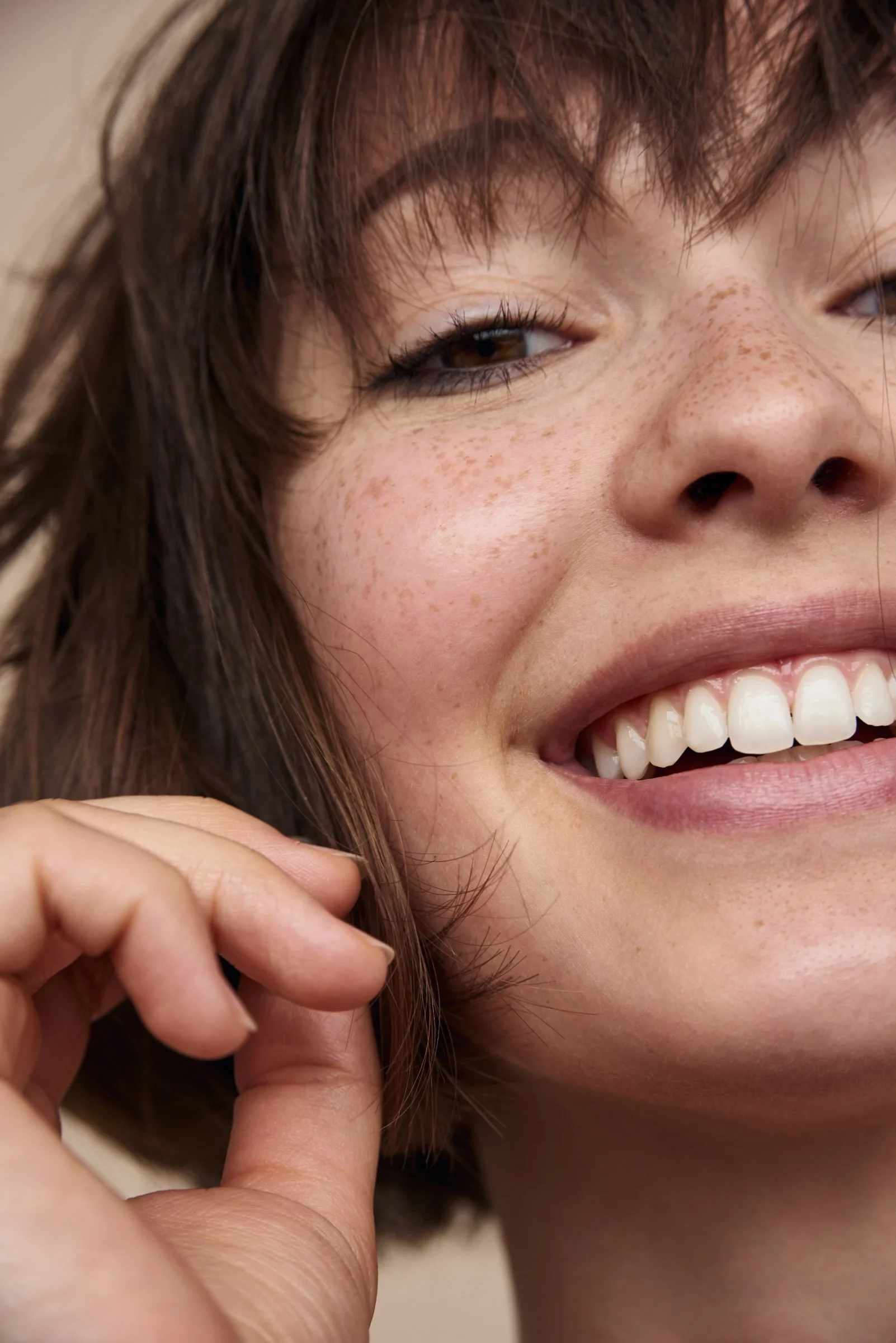 Close-up of a young woman smiling, showing teeth, with her hand near her face, and her brown hair with bangs.