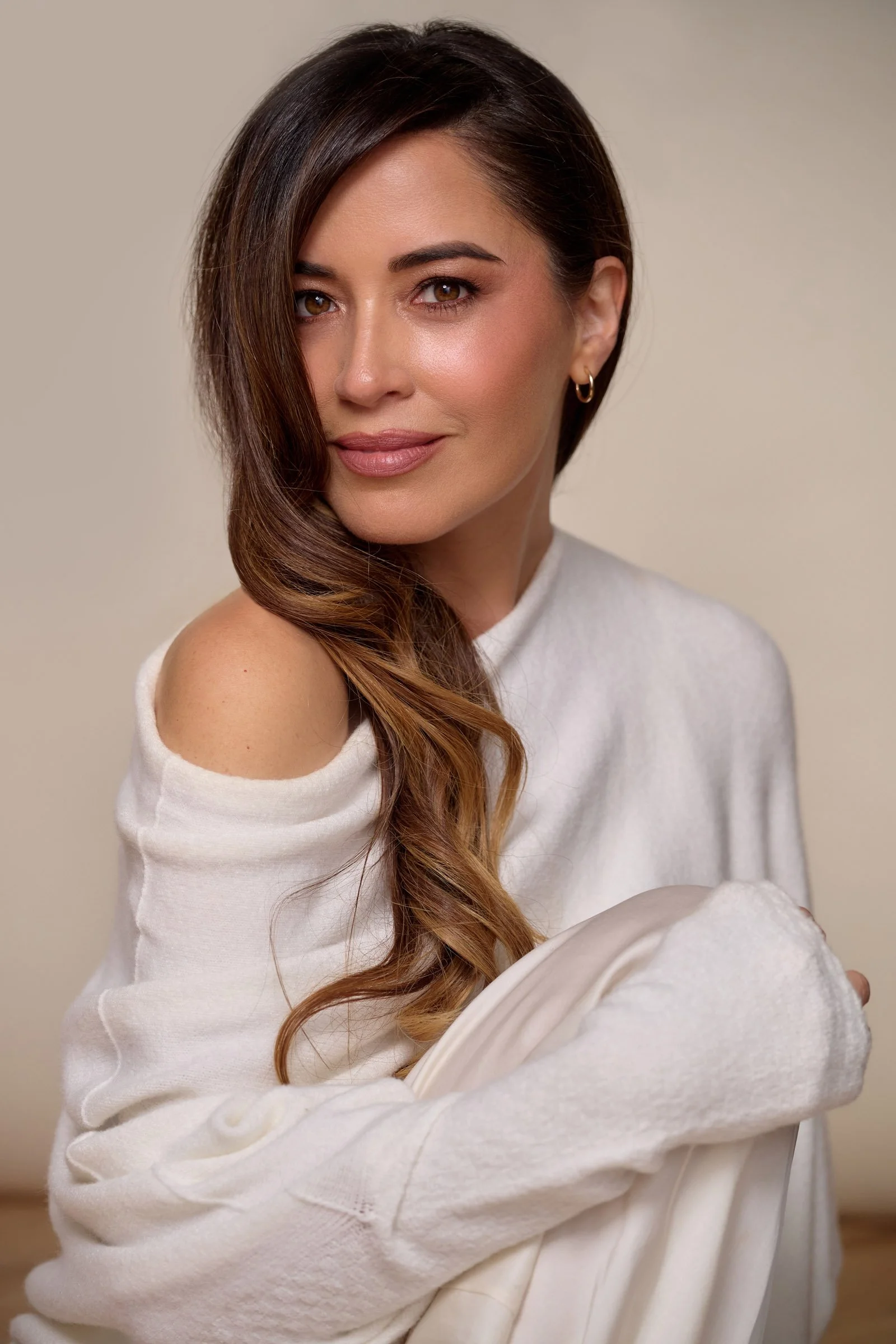 A woman with long wavy brown hair, wearing a white off-shoulder sweater, posing confidently against a neutral background.