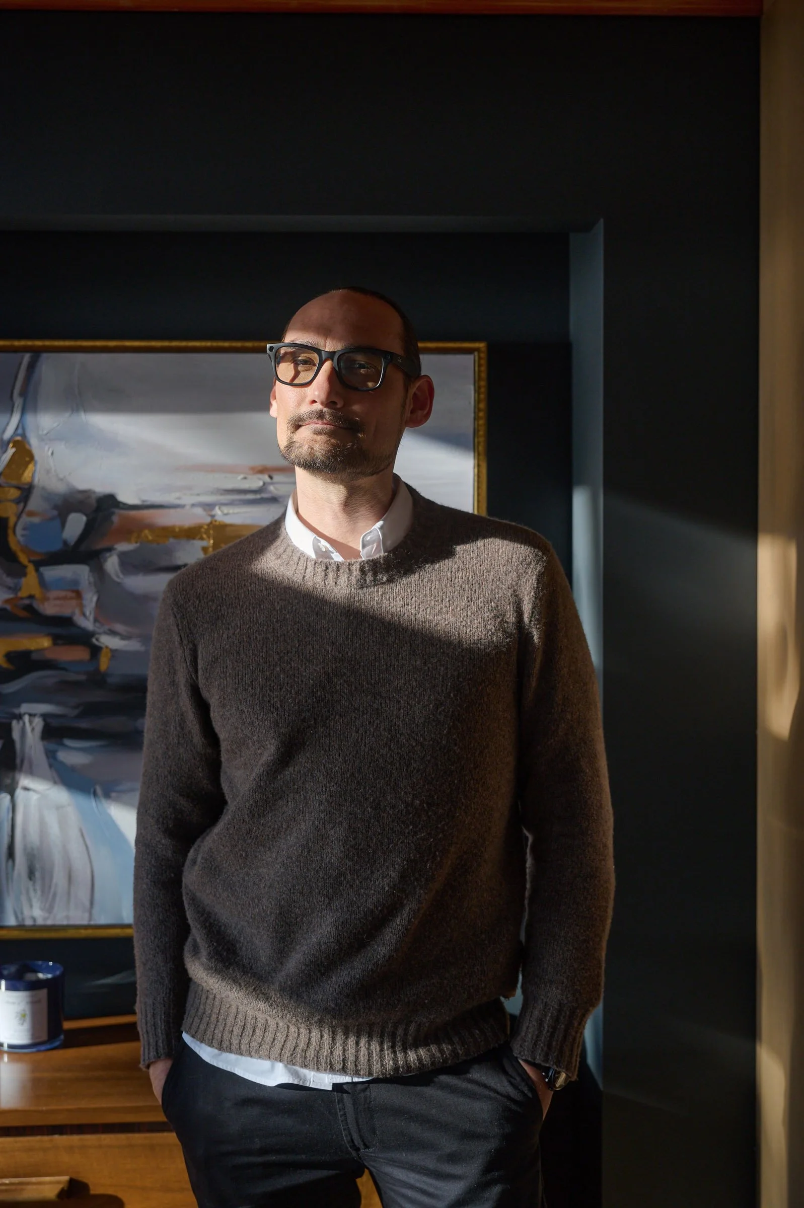 Man standing indoors wearing glasses, a brown sweater, white collared shirt, with hands in his pockets, in front of an abstract painting.