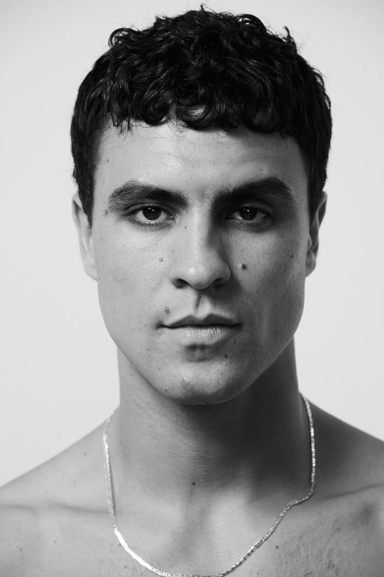 Black and white portrait of a young man with short, curly hair, gazing directly at the camera, with a necklace around his neck.