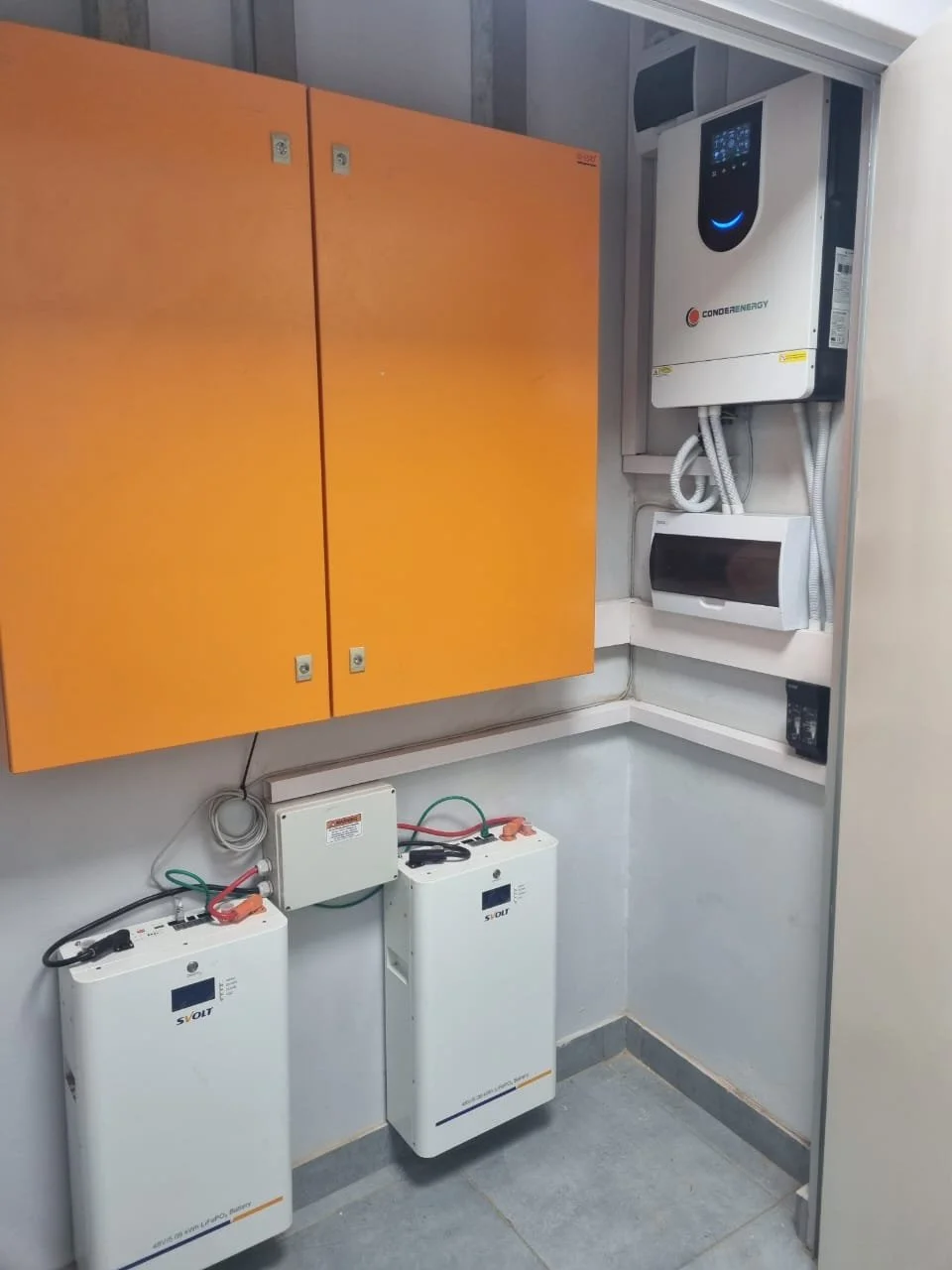 Electrical and energy storage equipment in utility room, including two batteries, a white inverter, and a white energy management system, with orange cabinets on the wall.