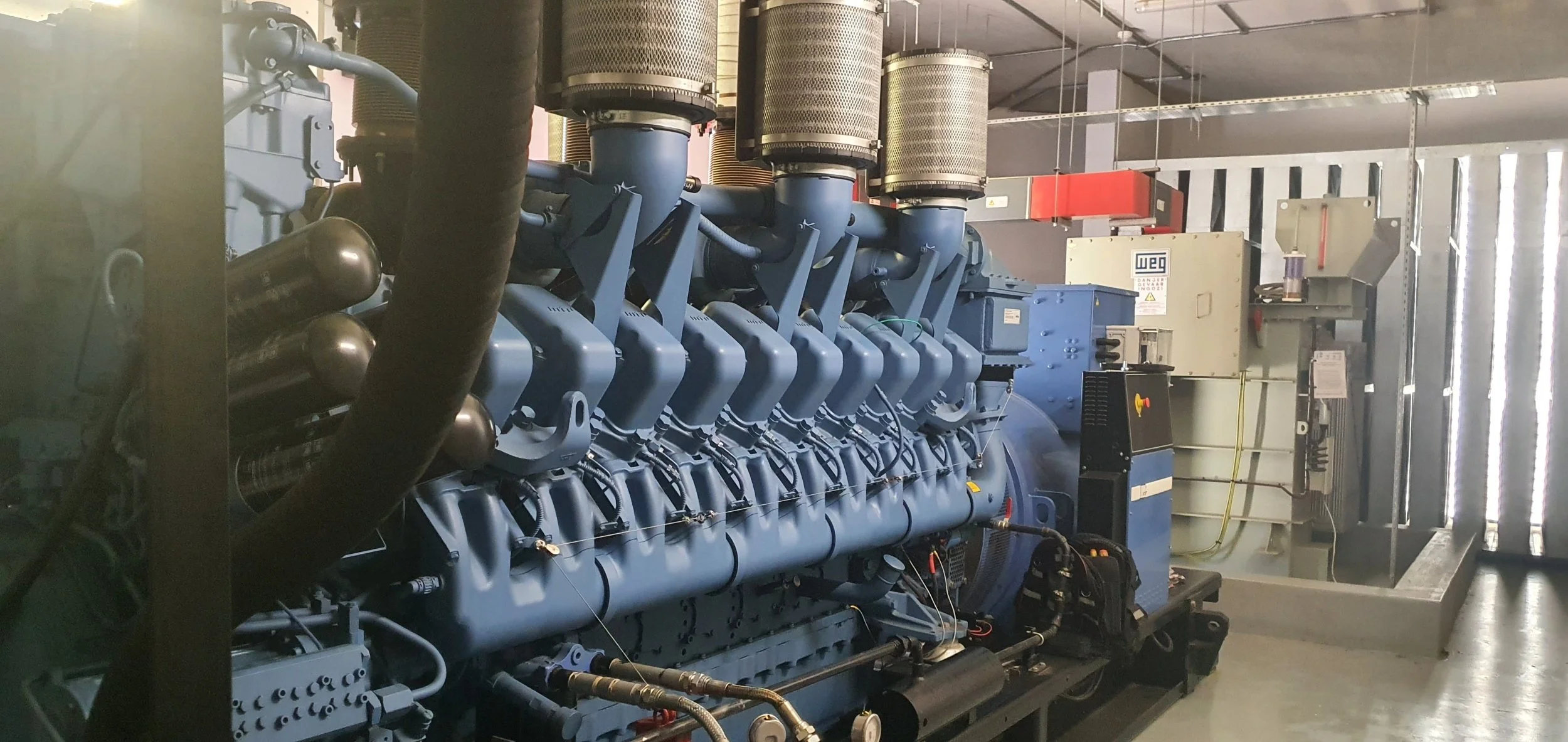 Large industrial blue generator or engine with multiple pipes and filters, situated in a factory or industrial setting.