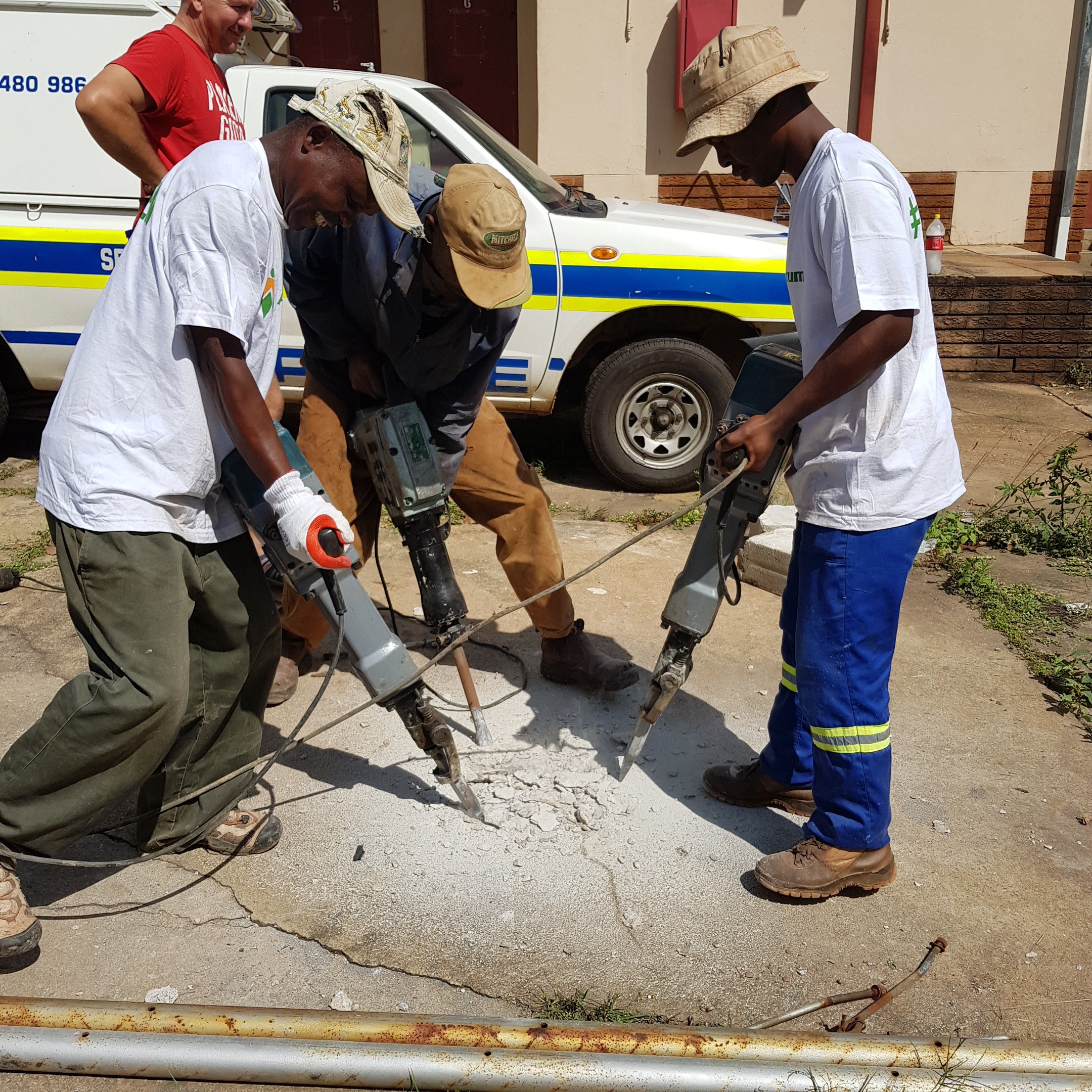 Three workers using jackhammers to break up concrete on a sidewalk, with a police vehicle and a man smiling in the background.