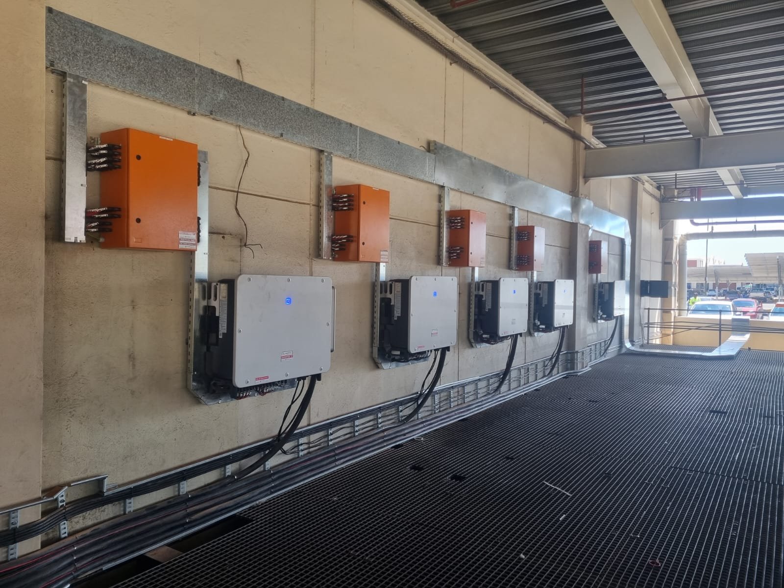 Multiple inverters or electrical control units installed on a wall outside, connected by cables and conduits, with a parking lot visible in the background.