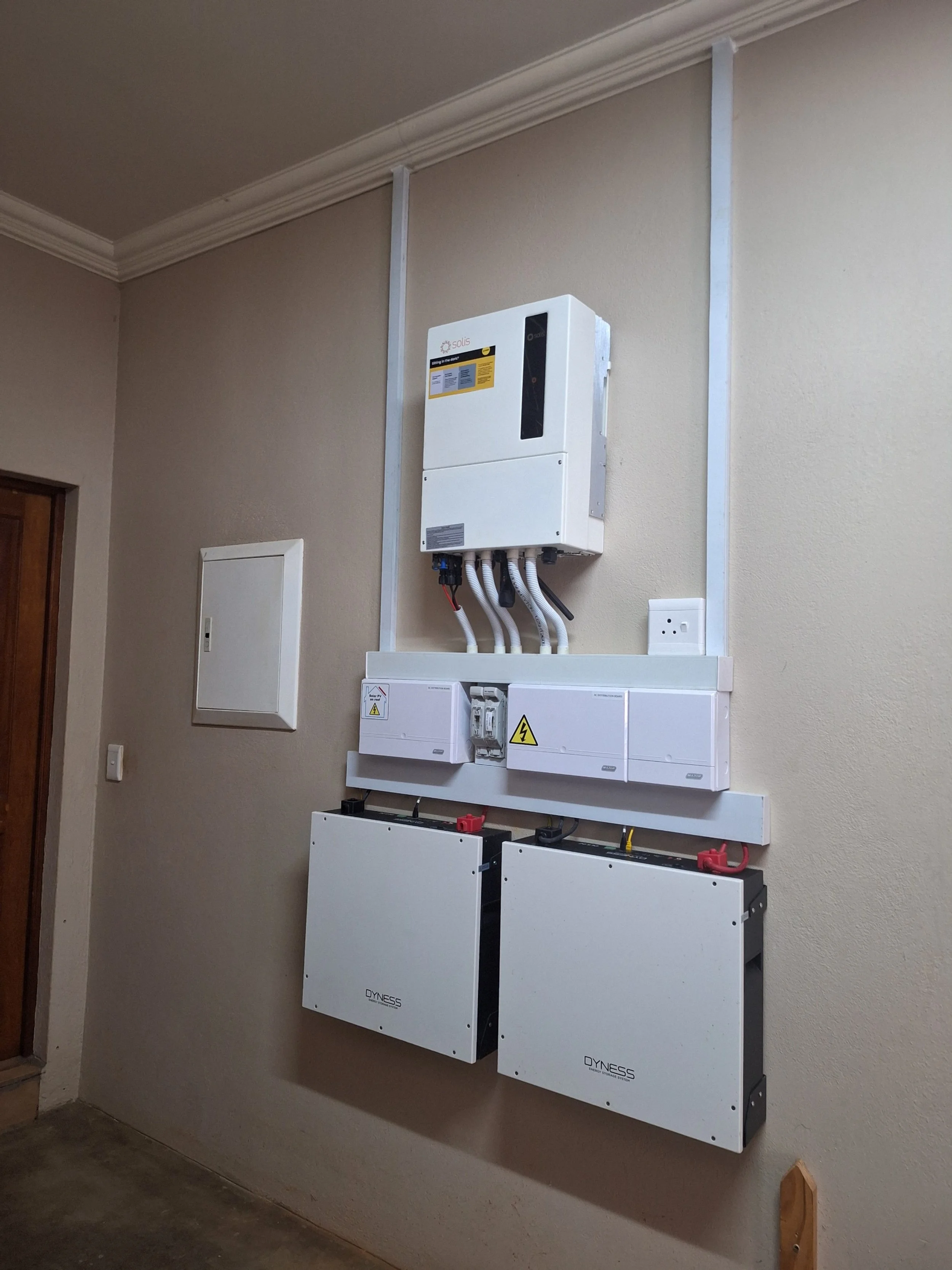 Electrical equipment installed on a beige wall in an indoor space, including an inverter, circuit breaker panels, and power distribution units.