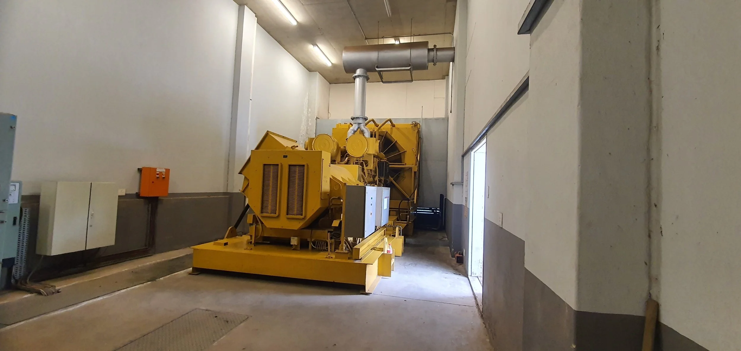 Large yellow industrial machine in a warehouse, connected to an exhaust duct, with control panels and electrical boxes nearby.