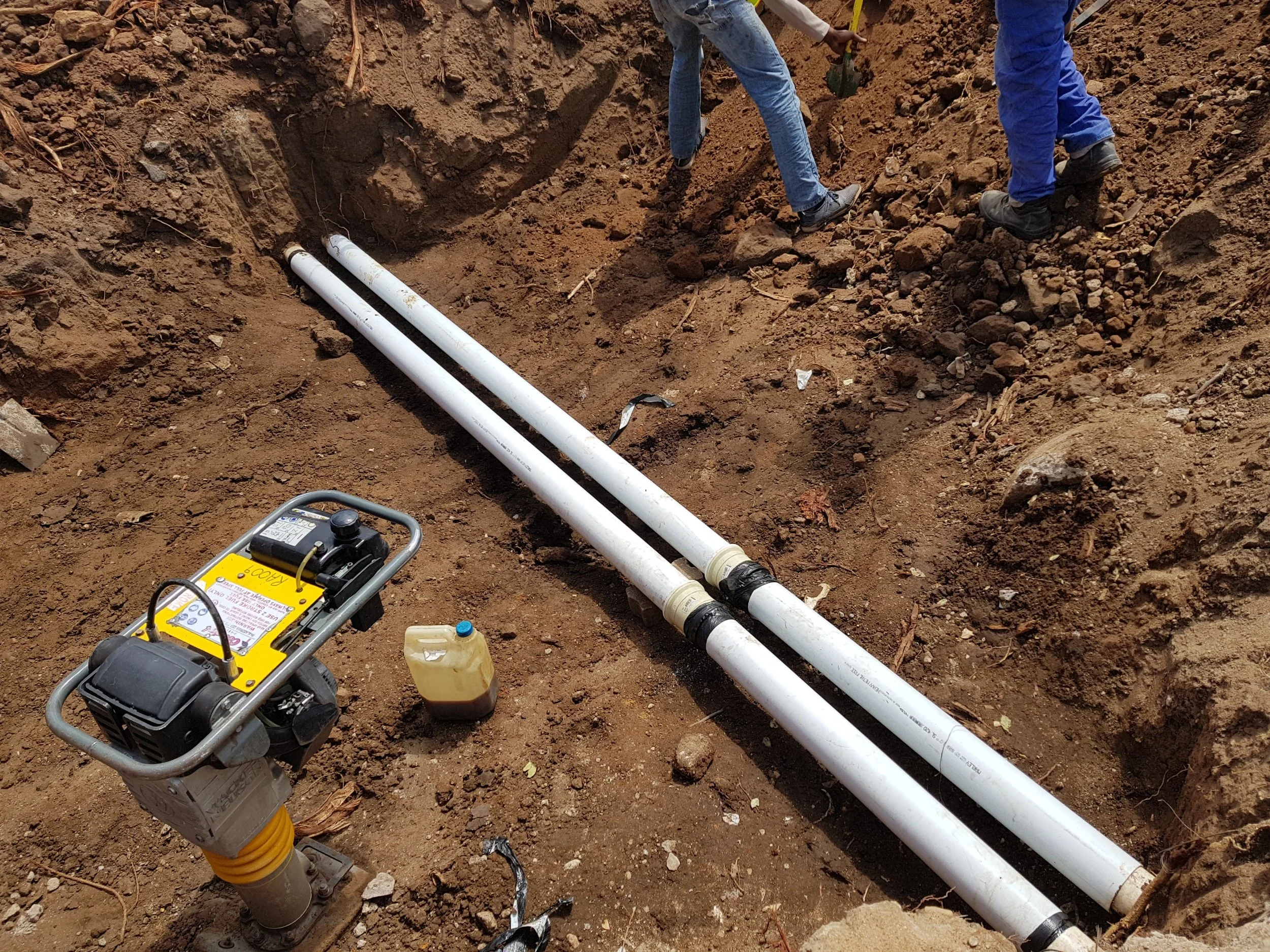 Two workers are installing or repairing underground pipes with two white PVC pipes in a trench, along with a base machine and a yellow liquid container nearby.