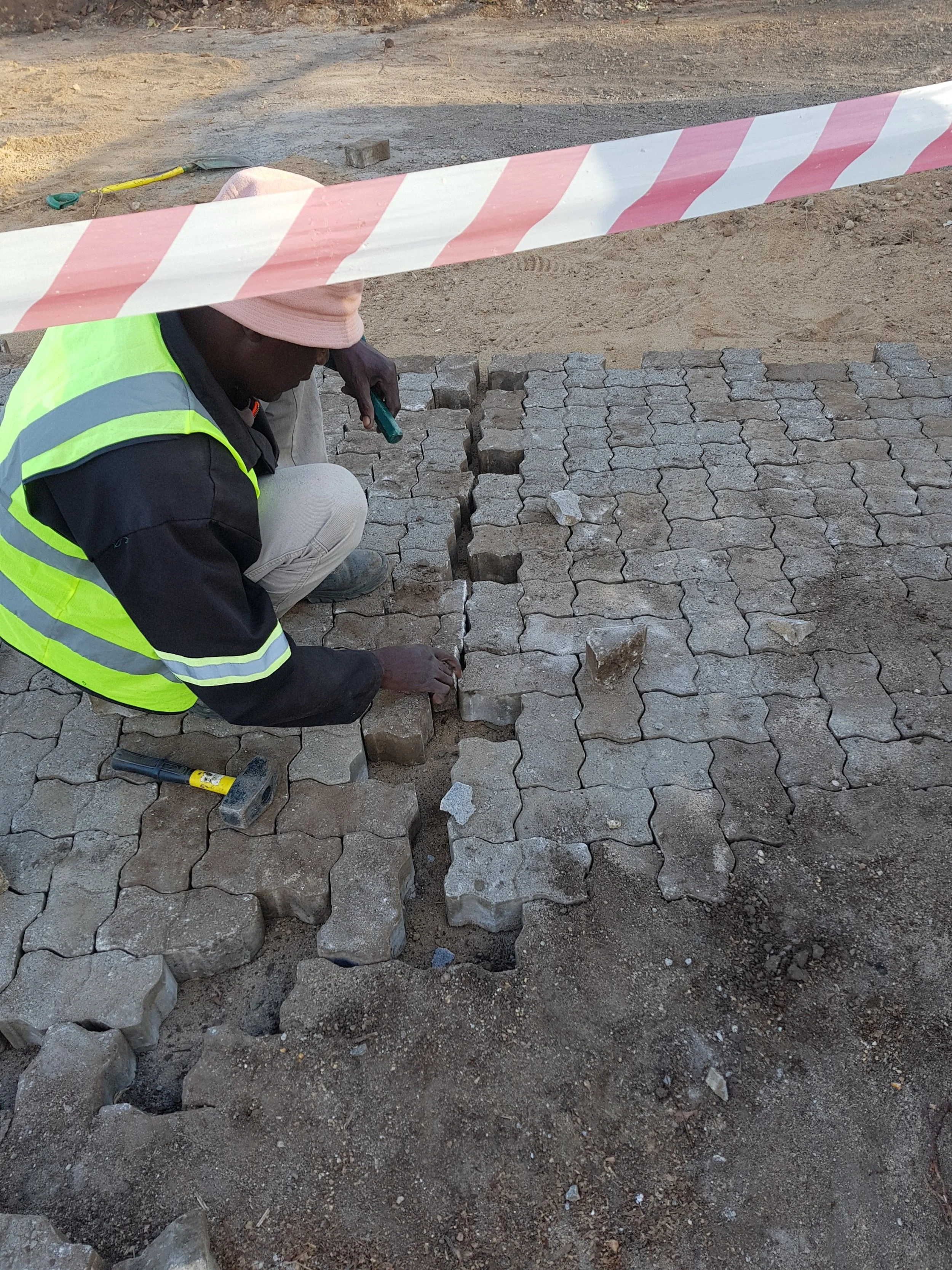 Worker laying paving stones on a construction site with safety tape overhead.