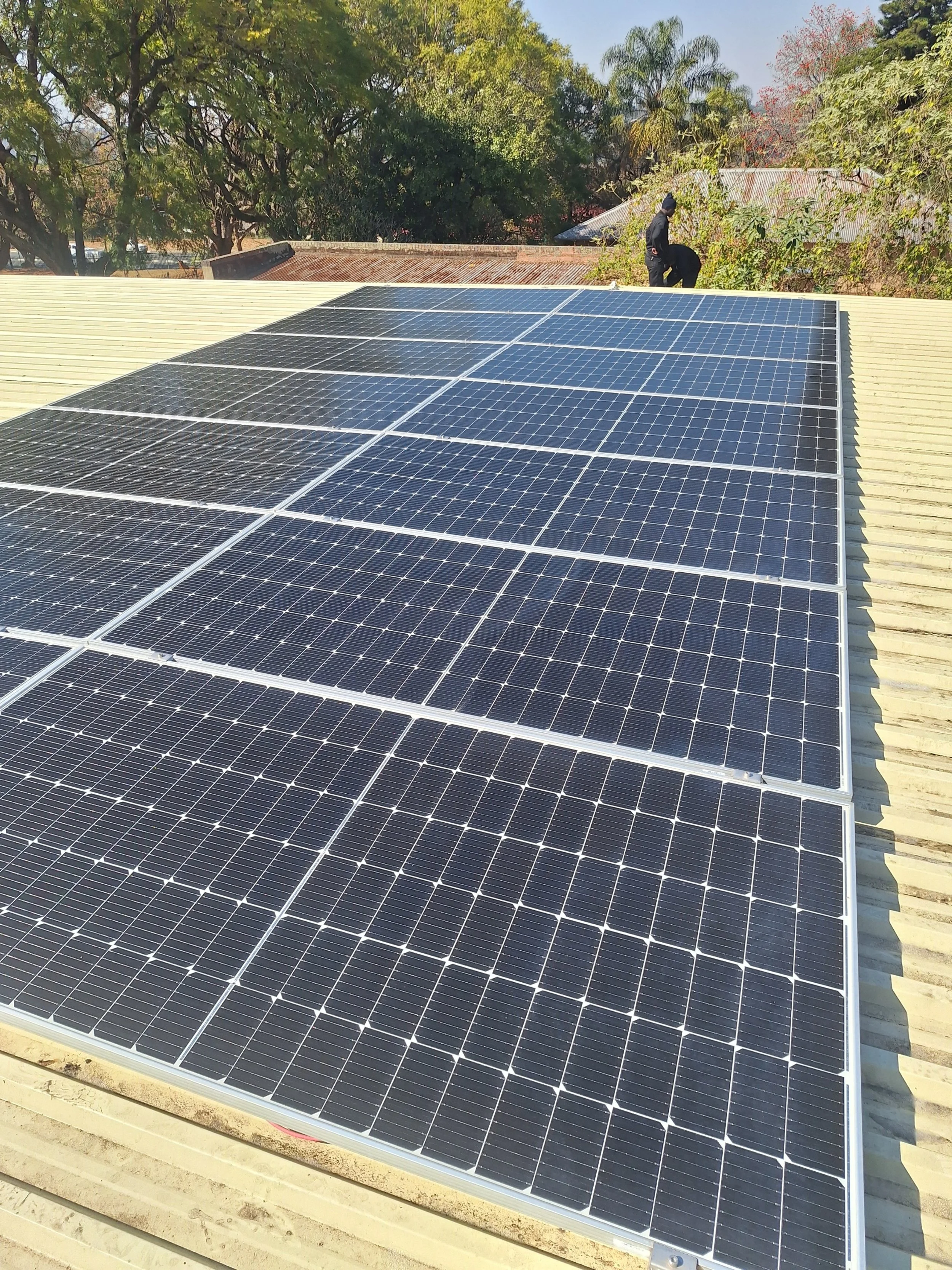 Close-up of solar panels installed on a roof, with a person working in the background among trees and buildings.