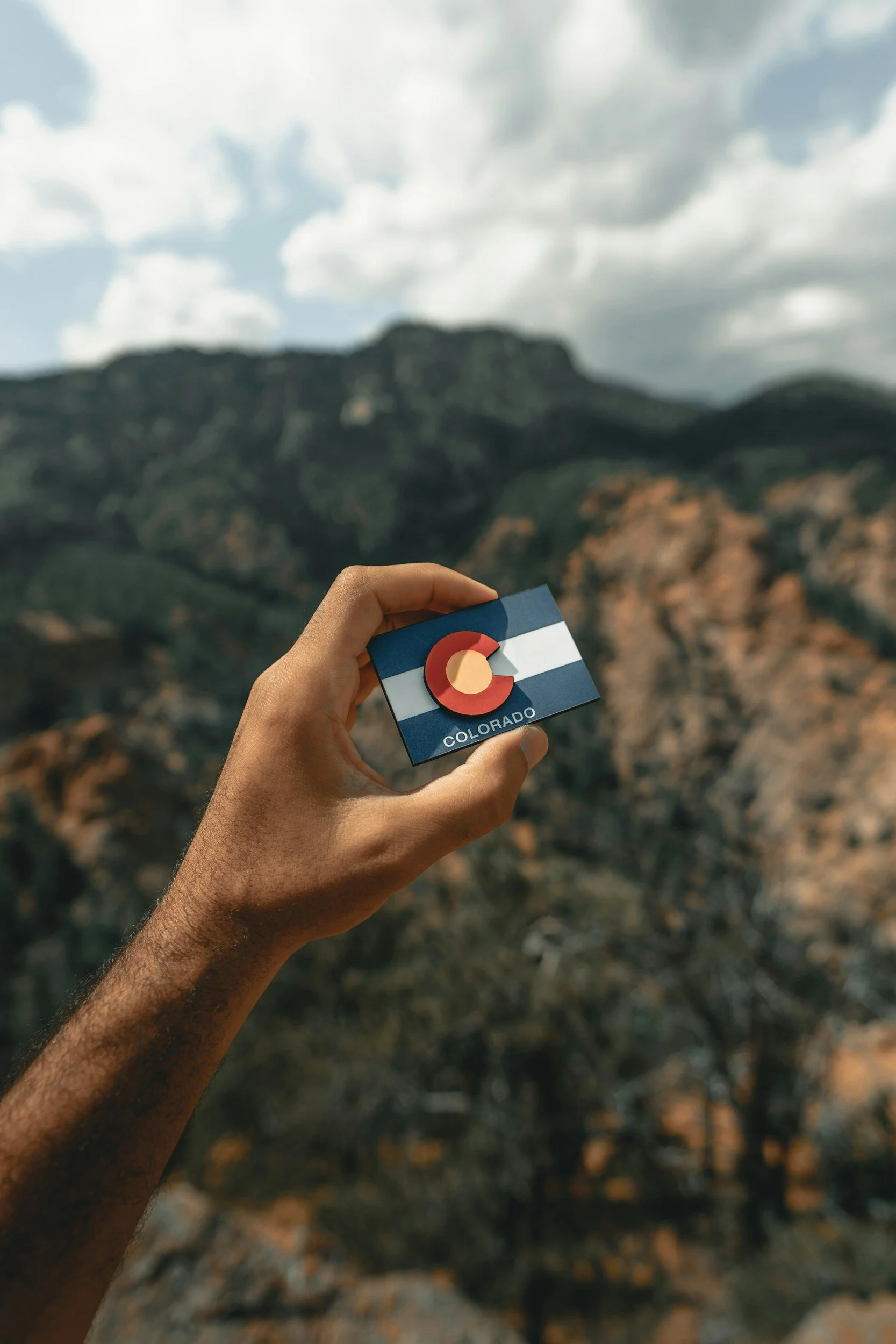 Person holding a Colorado state card against a mountain landscape with cloudy sky.