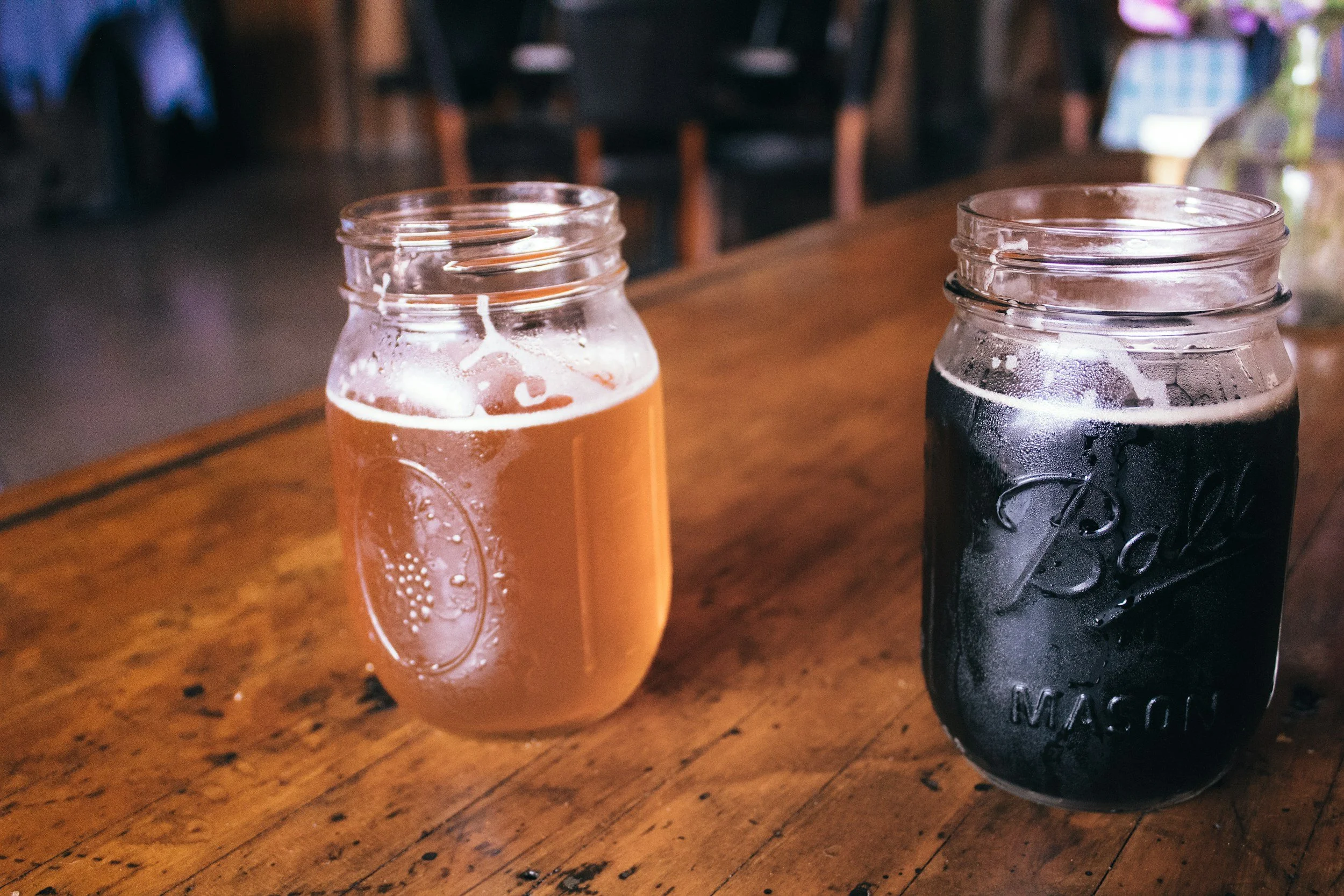 Two glass jars with drinks on a wooden table, one with a light-colored beverage and the other with a dark beverage, both with condensation on the outside.