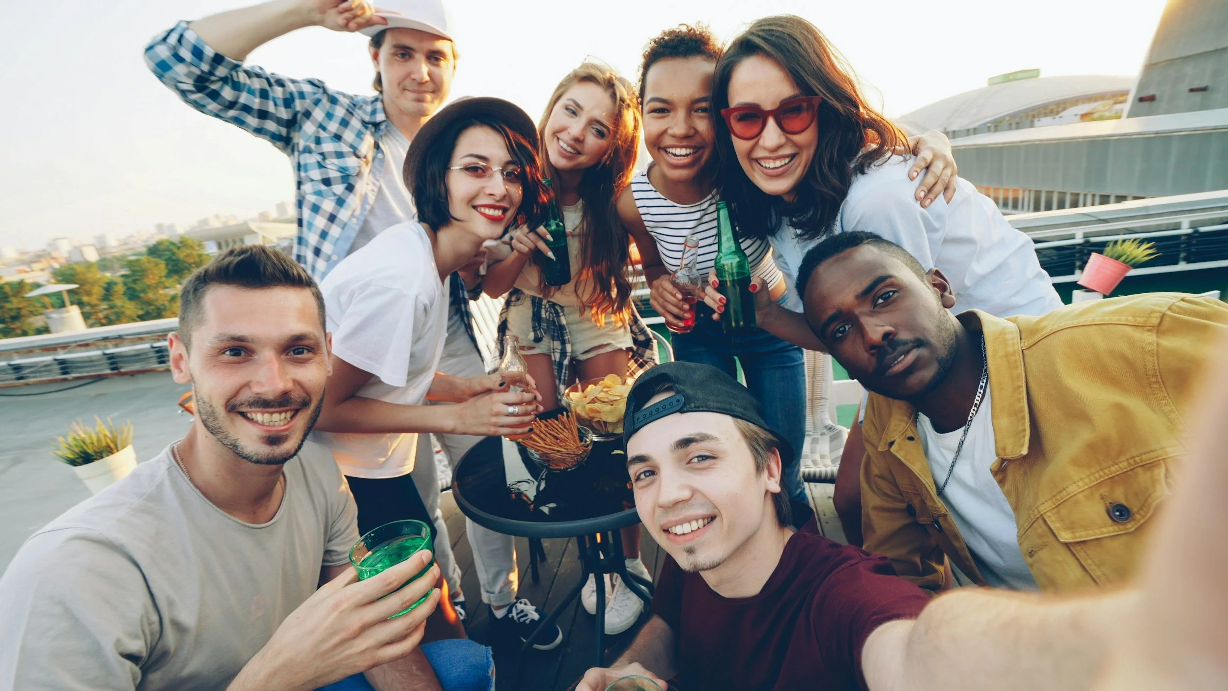 Group of friends smiling and taking a selfie on a rooftop during sunset, with some holding drinks and snacks, enjoying a casual gathering.