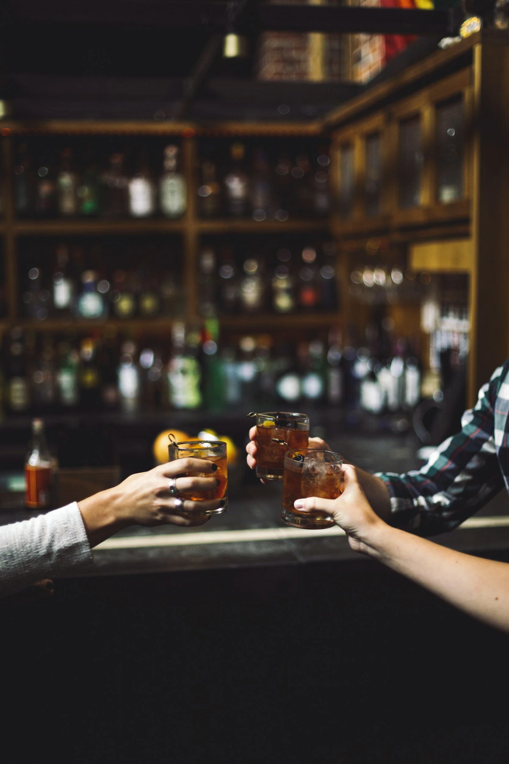 Two people clinking glasses with drinks at a bar, with a wooden shelf filled with bottles in the background.