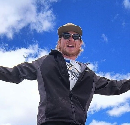 A man wearing sunglasses, a cap, and a black jacket is outdoors with arms outstretched against a blue sky with clouds.