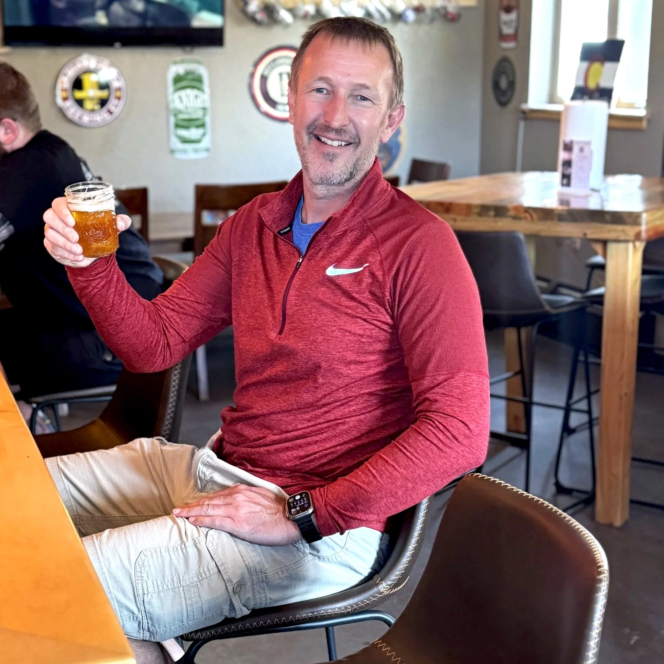 A smiling man in a red long-sleeve Nike shirt, beige shorts, and a smartwatch, sitting in a casual restaurant or brewery, holding a glass of beer, with a light-filled interior and decorative wall signs in the background.