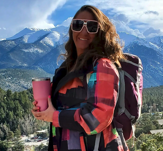 Woman wearing sunglasses and a colorful jacket, carrying a backpack, holding a pink travel mug, standing outdoors with snow-capped mountains and greenery in the background.