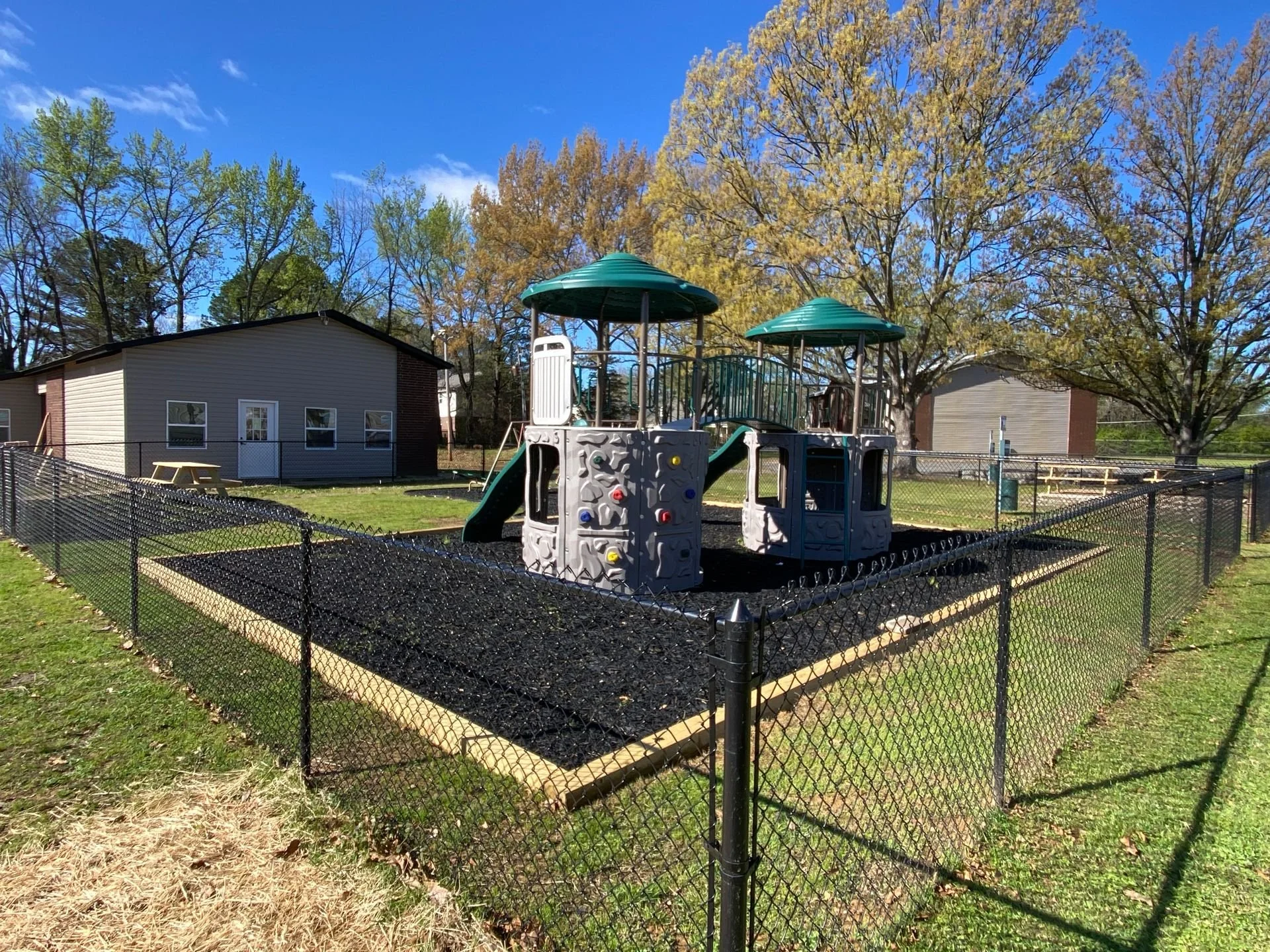 Children's playground with slides and climbing structure, enclosed by black chain-link fence, on a grassy area with trees and buildings in the background under a blue sky.