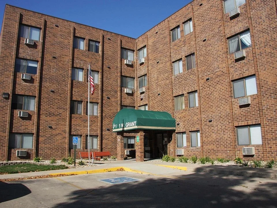 Brick apartment building with a green awning labeled 1 W. Grant, parking lot with handicap parking space, American flag, and a bench in front of the building.