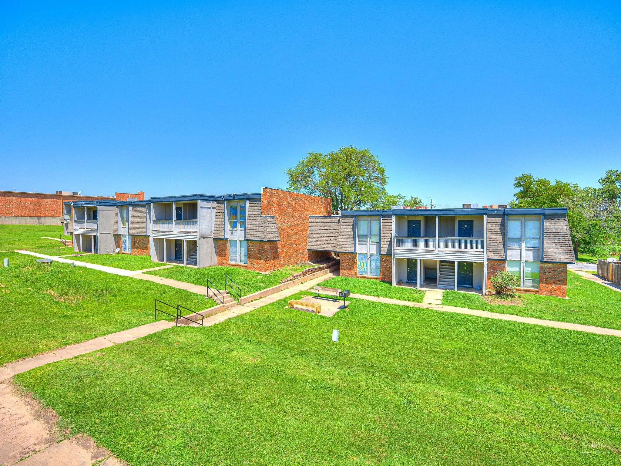 Exterior of a two-story apartment complex with brick and wood siding, multiple staircases, walkways, and green lawn under a bright blue sky.