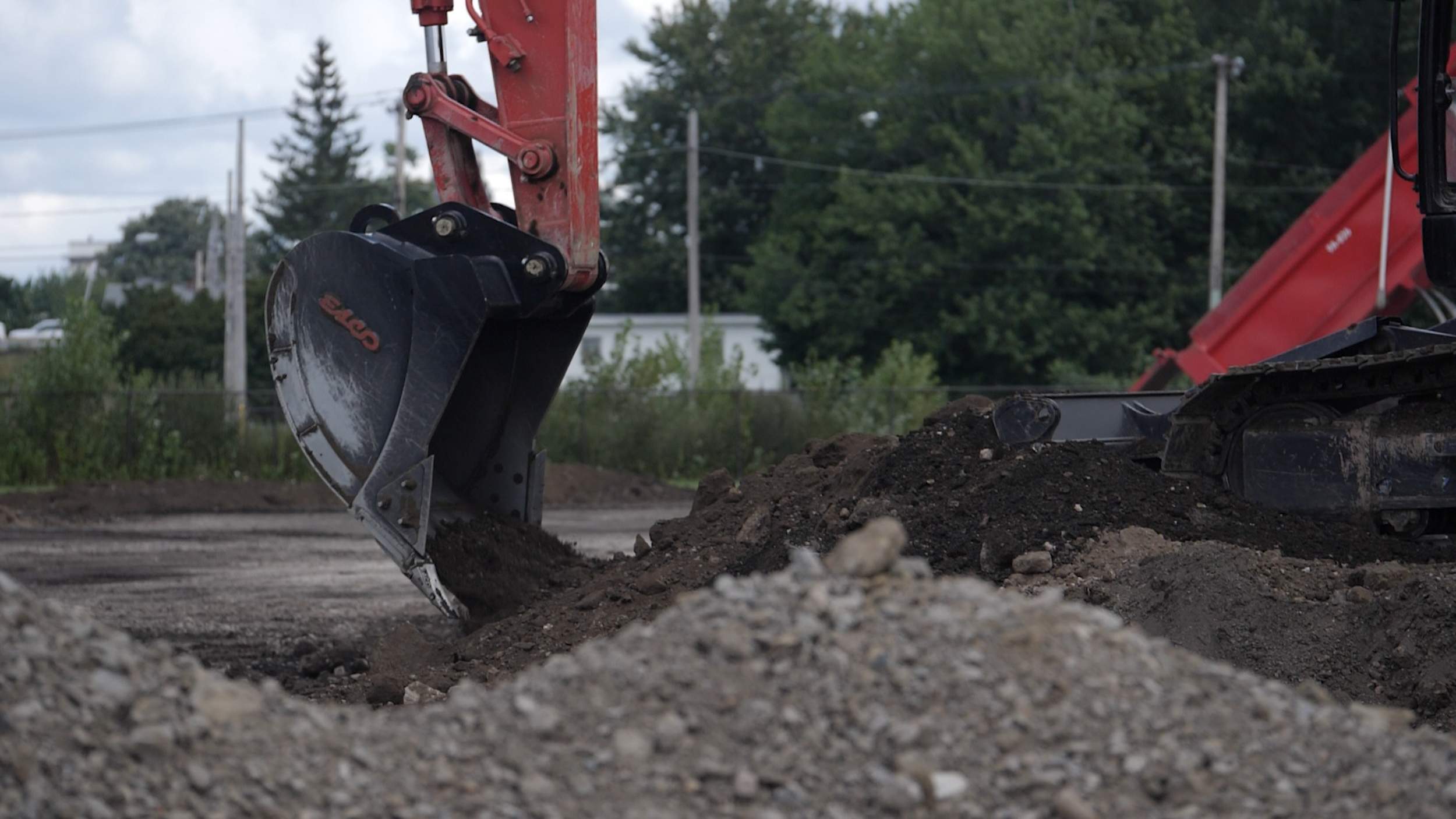 Close-up of an excavator bucket digging into dirt on a construction site, with trees and utility poles in the background.