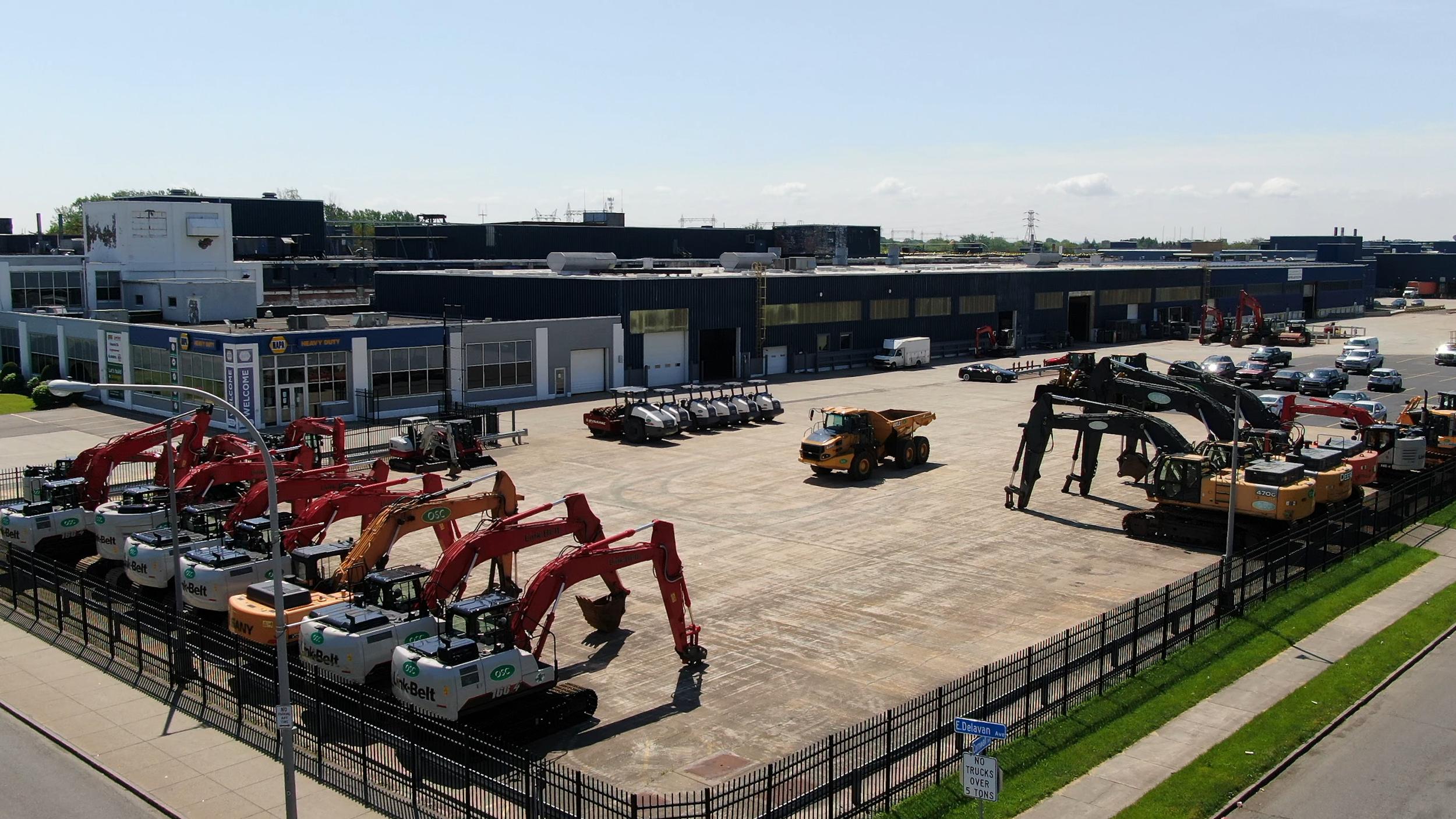 A large industrial equipment yard with construction machinery, trucks, and parked vehicles outside a warehouse building, surrounded by a black fence. There are multiple excavators in the foreground, other construction vehicles, and a parking lot with cars visible on the right.