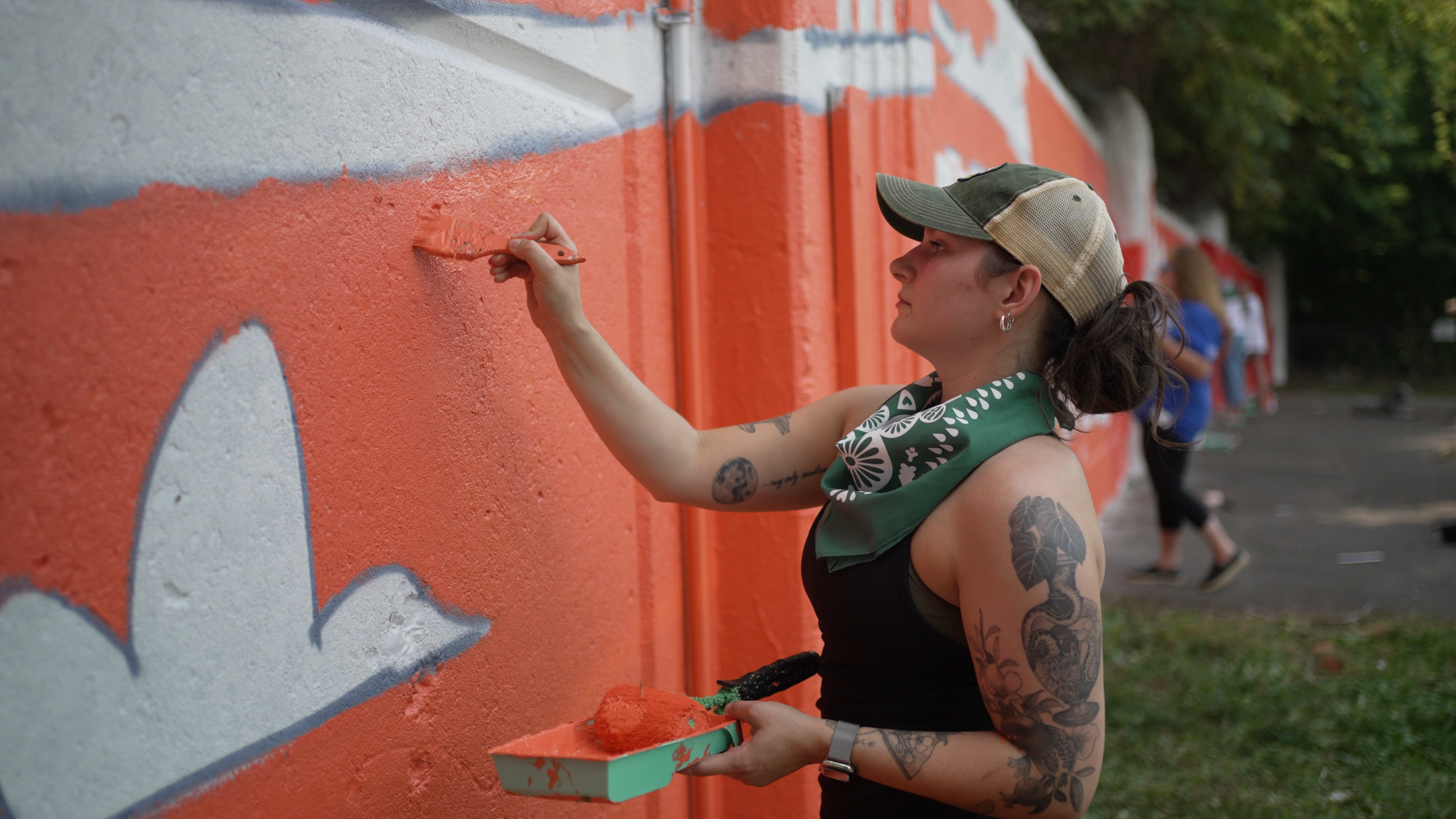 A woman with tattoos on her arms and neck painting orange on a graffiti wall with a roller brush while holding a paint tray.