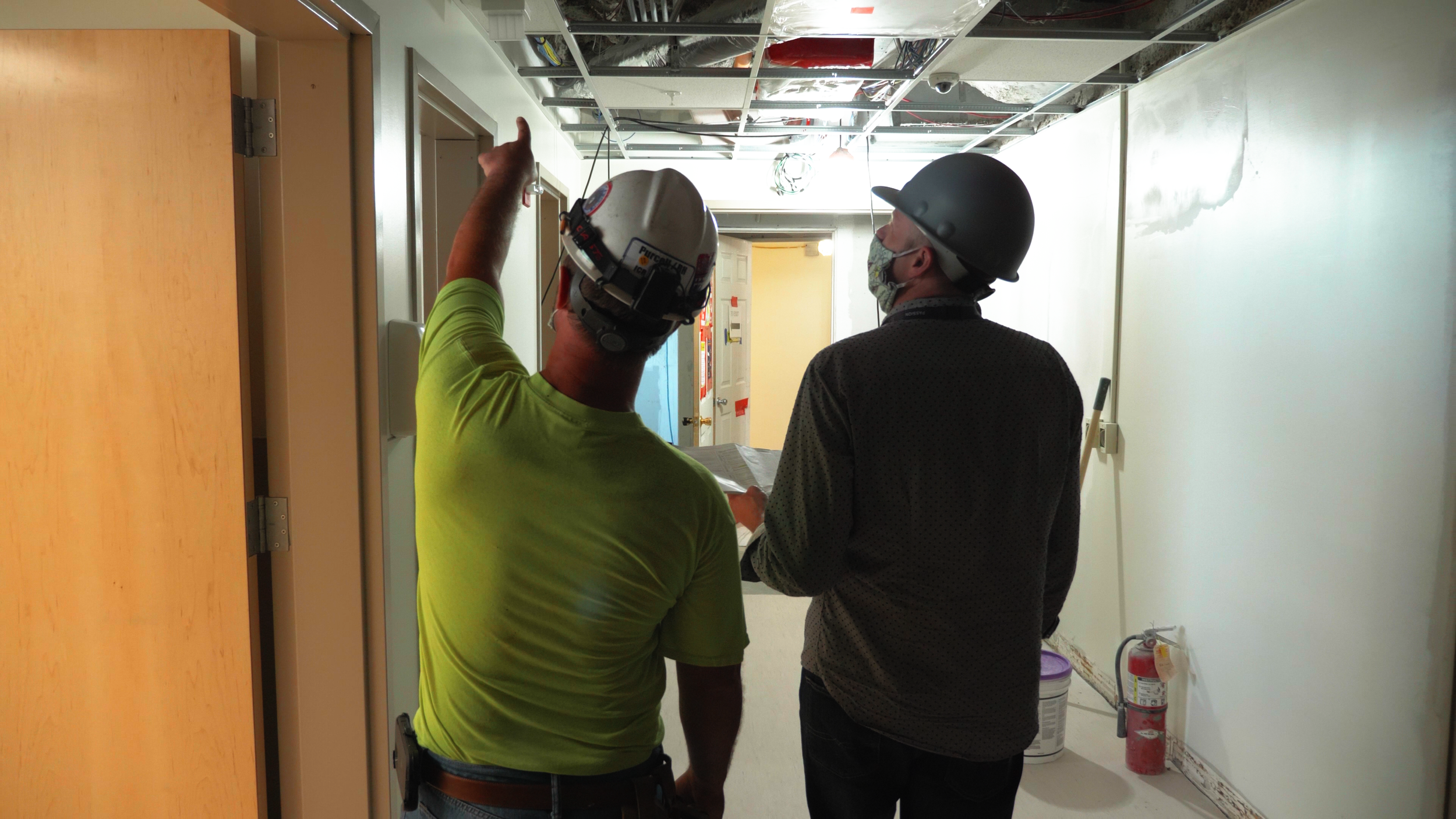 Two construction workers, wearing helmets and masks, are discussing plans in a hallway with exposed ceiling wires and an open ceiling panel. One worker points at the ceiling.