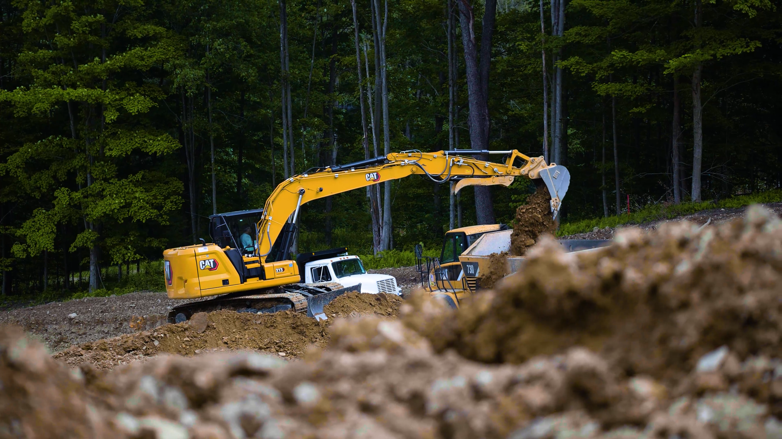 Yellow excavator moving dirt on a construction site near a wooded area.